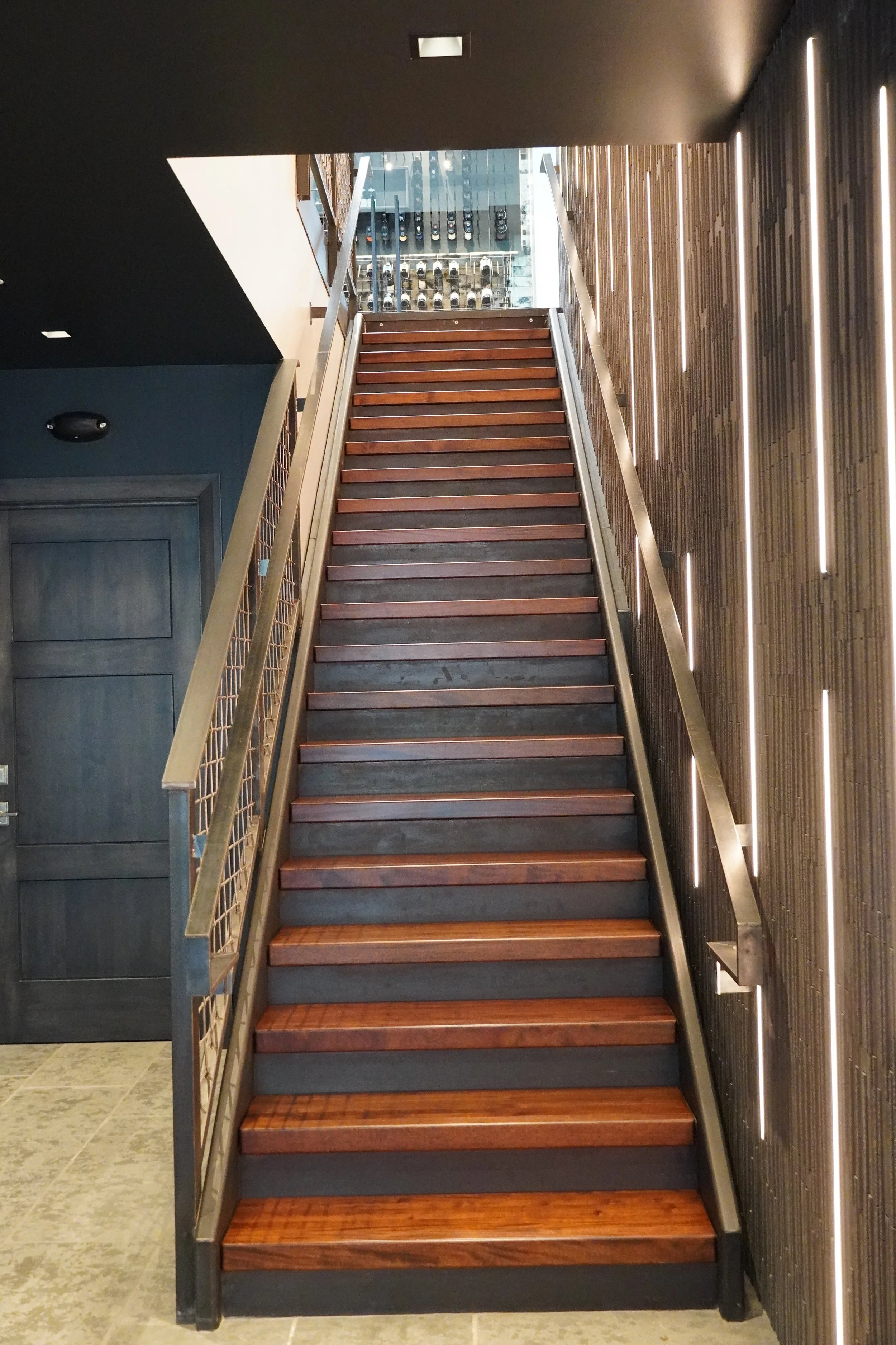 A staircase with wooden steps and metal railings leading upwards, with a glass wall revealing parked cars outside.