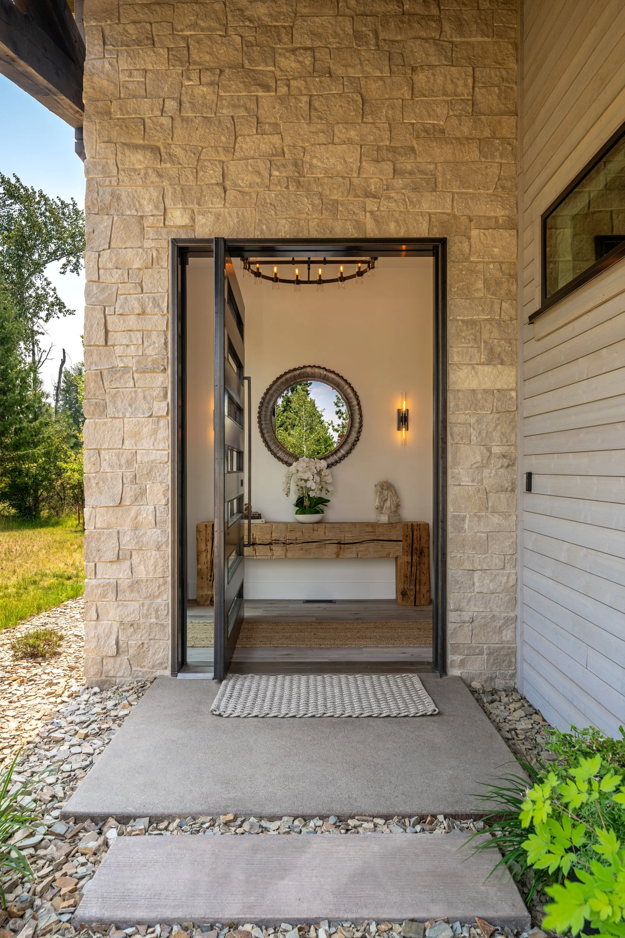 Front door opening into a modern entryway with a stone and wood exterior, a mirror, a chandelier, a vase with white flowers, and decorative art on a wooden console table.