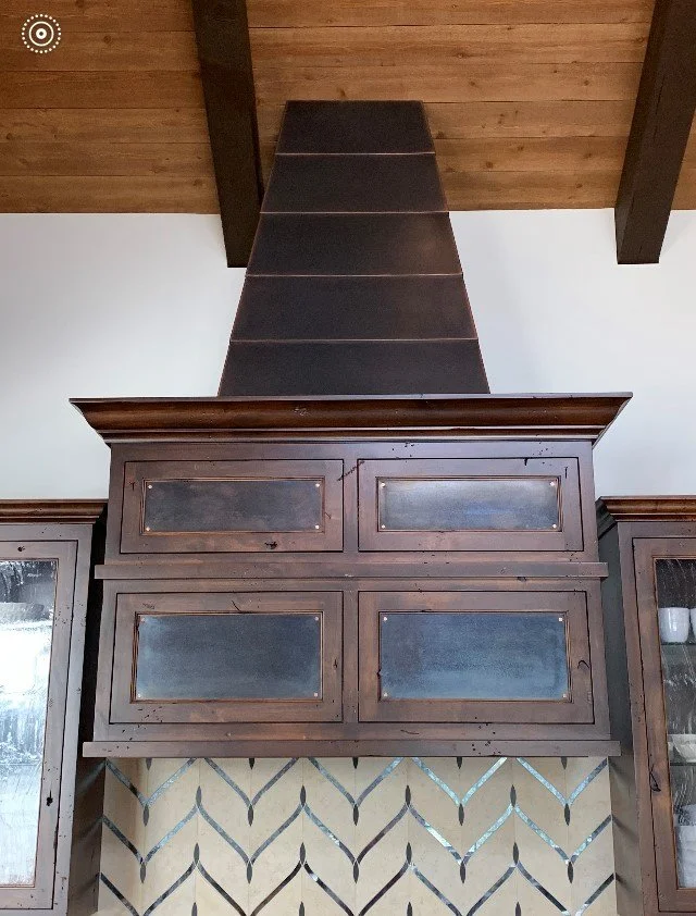 View of a kitchen range hood made of dark wood with glass-front cabinets on either side and a decorative backsplash below.