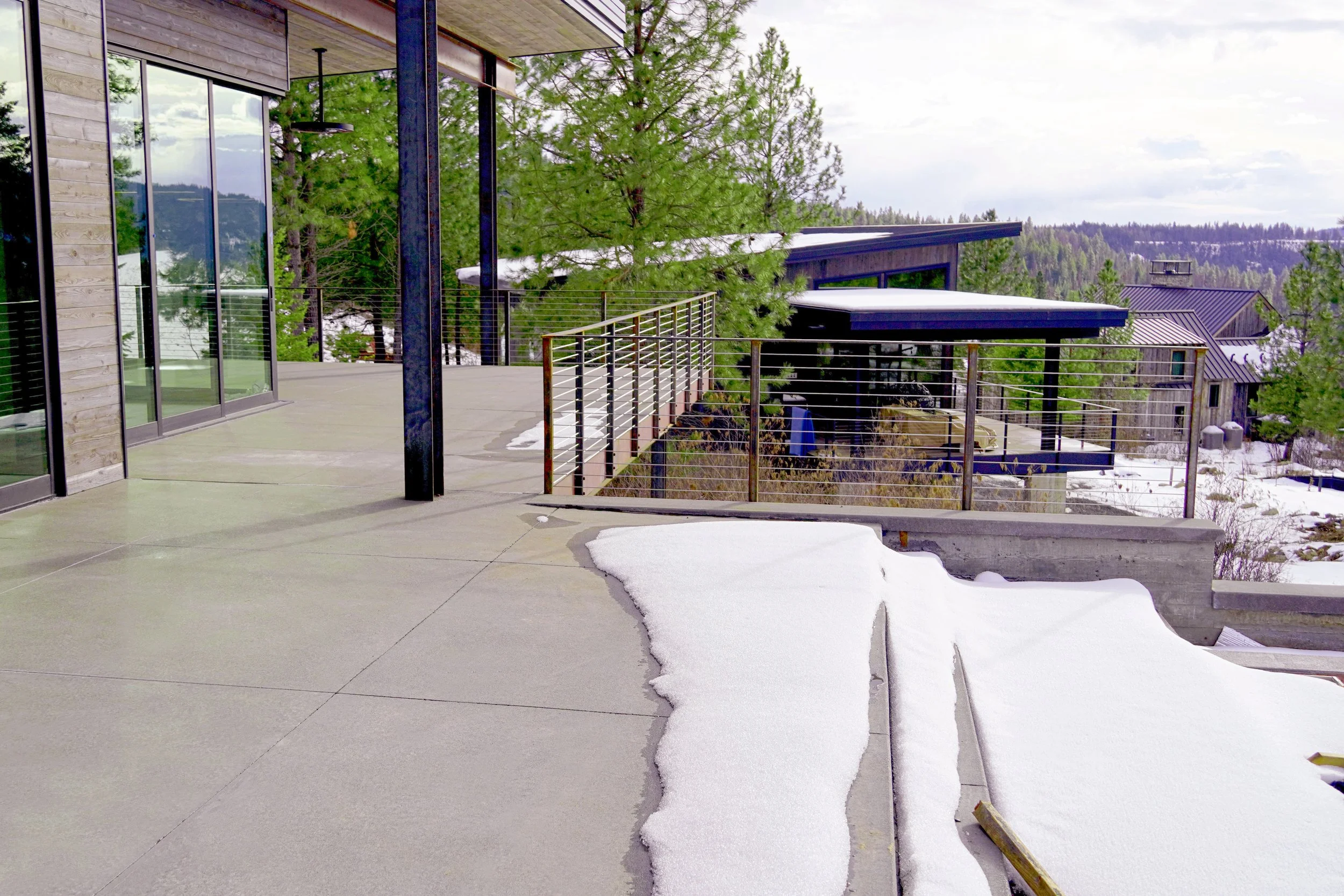 A modern balcony with glass doors, concrete flooring, and a metal railing overlooking a snowy landscape with trees and houses.