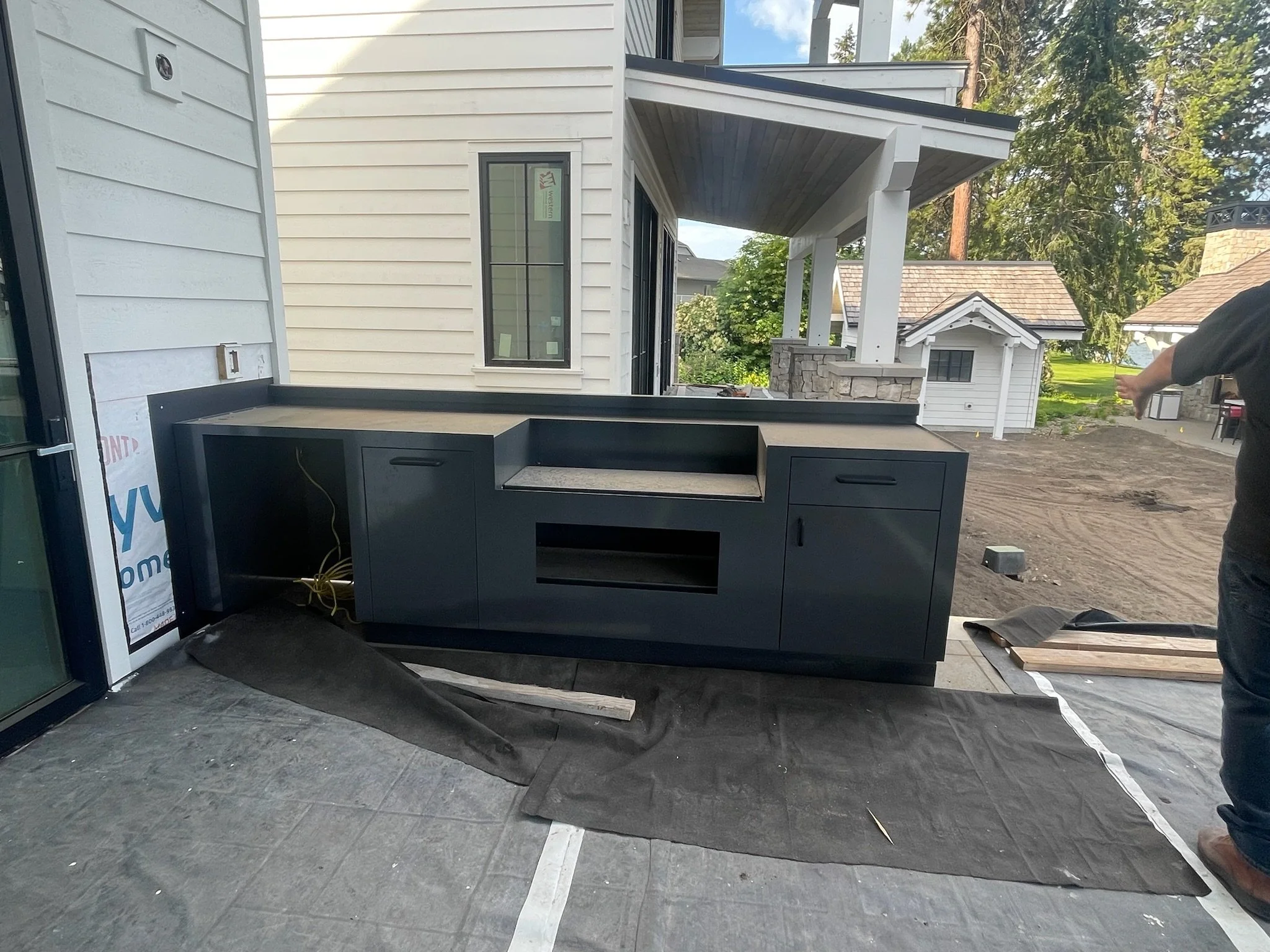 Outdoor kitchen or BBQ unit under construction with black cabinetry and granite countertop, on a patio with black fabric covering the ground, adjacent to a white house with siding, in a backyard with trees and other structures visible in the backgrou