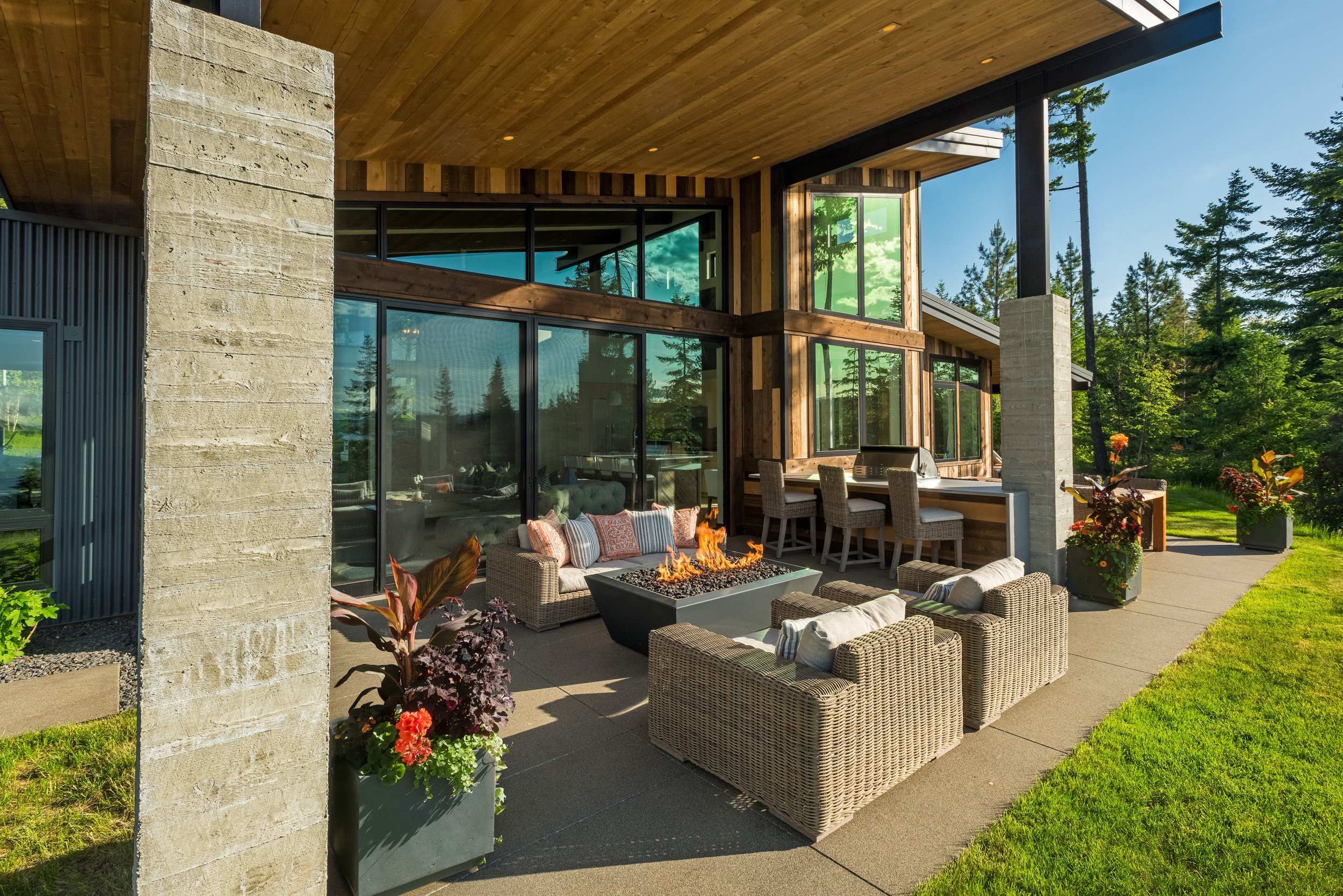 Exterior view of a modern house with a spacious patio area featuring outdoor furniture, a fire pit, and potted plants, surrounded by green grass and trees.