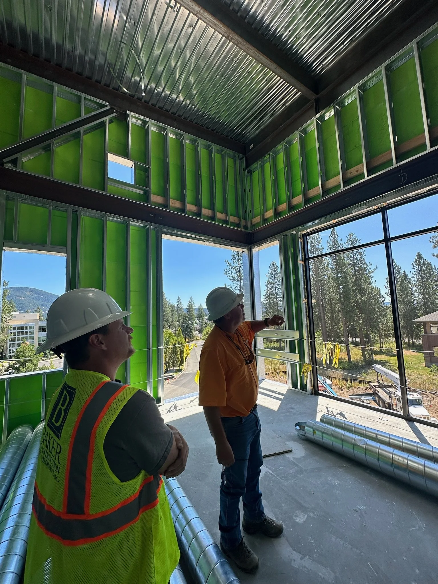 Two construction workers wearing safety helmets inside a building under construction with insulated green wall panels and large windows showing an outdoor scene with trees and a clear sky.