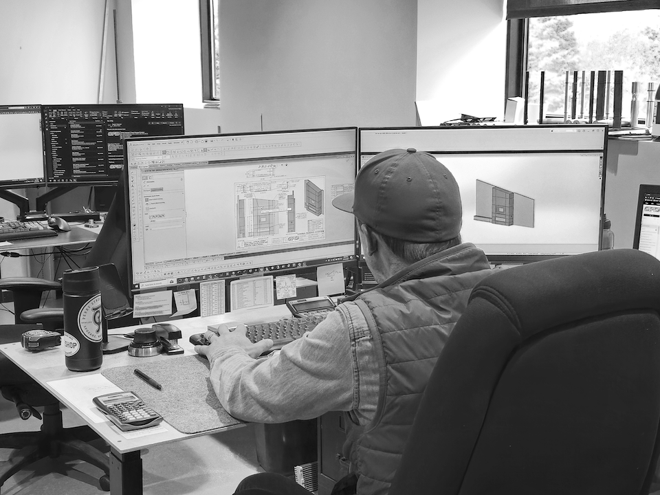 Person working on a CAD design at a computer desk with multiple monitors in an office.
