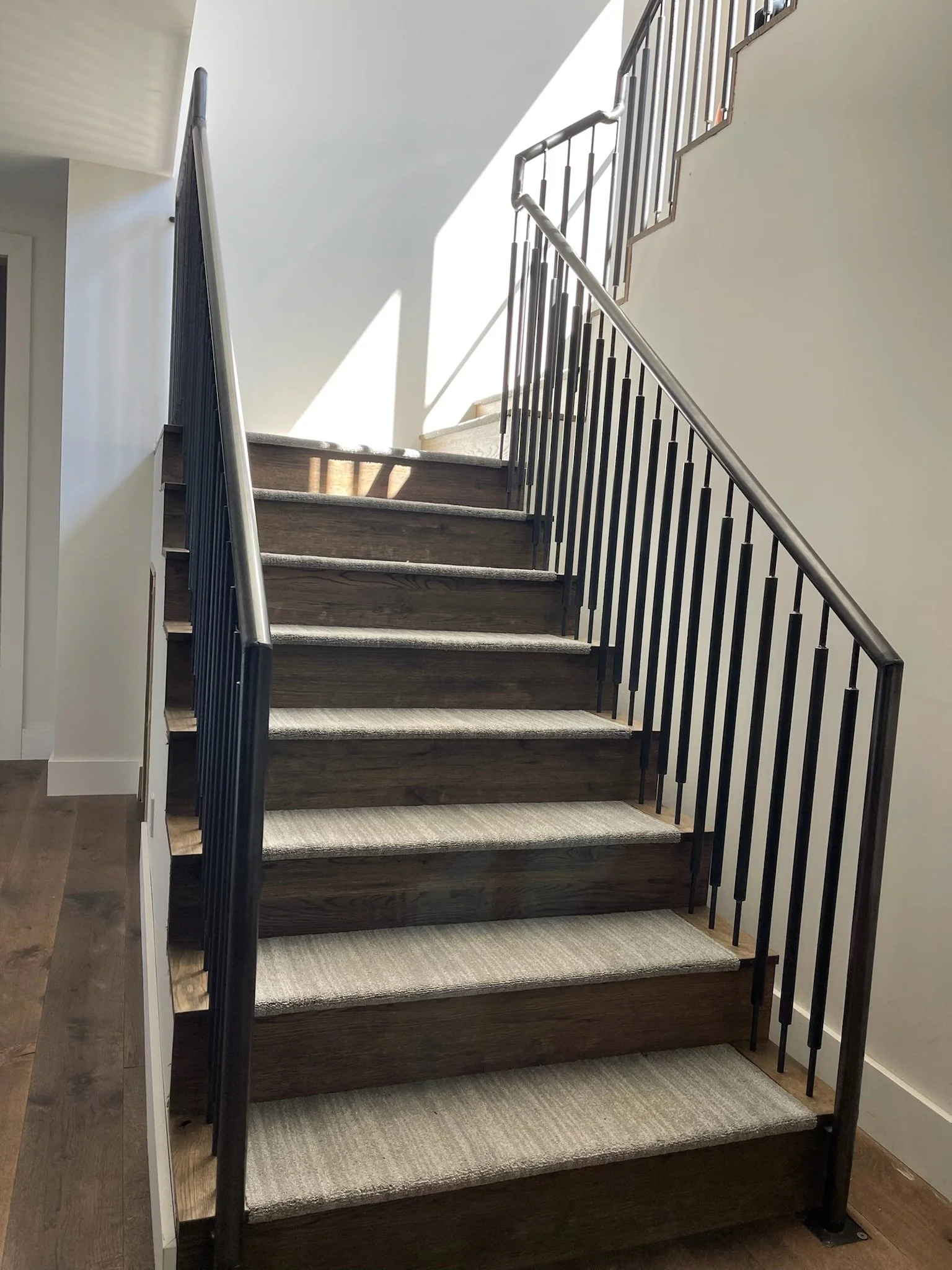 Indoor staircase with wooden steps, black metal railings, and a carpet runner, leading upwards with sunlight casting shadows.