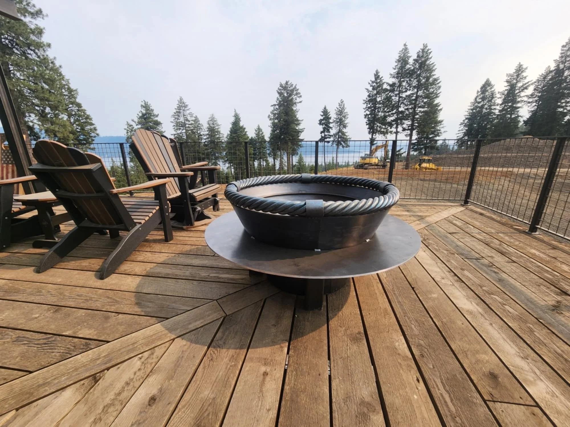 Outdoor wooden deck with a round fire pit and four wooden Adirondack chairs, surrounded by a metal fence, with pine trees and construction equipment in the background.