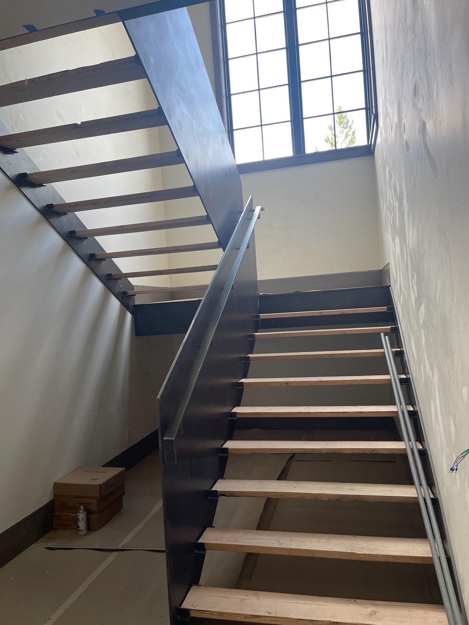 Interior view of a staircase under construction with wooden steps, metal railing, and a large window allowing natural light in.
