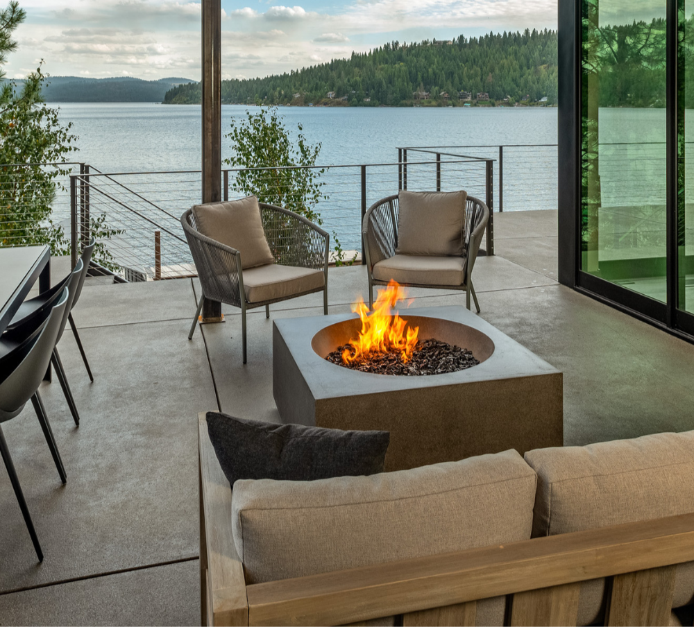 Contemporary outdoor patio with a fire pit, two cushioned chairs, a sofa, dining chairs, and a balcony overlooking a lake and forested hills.