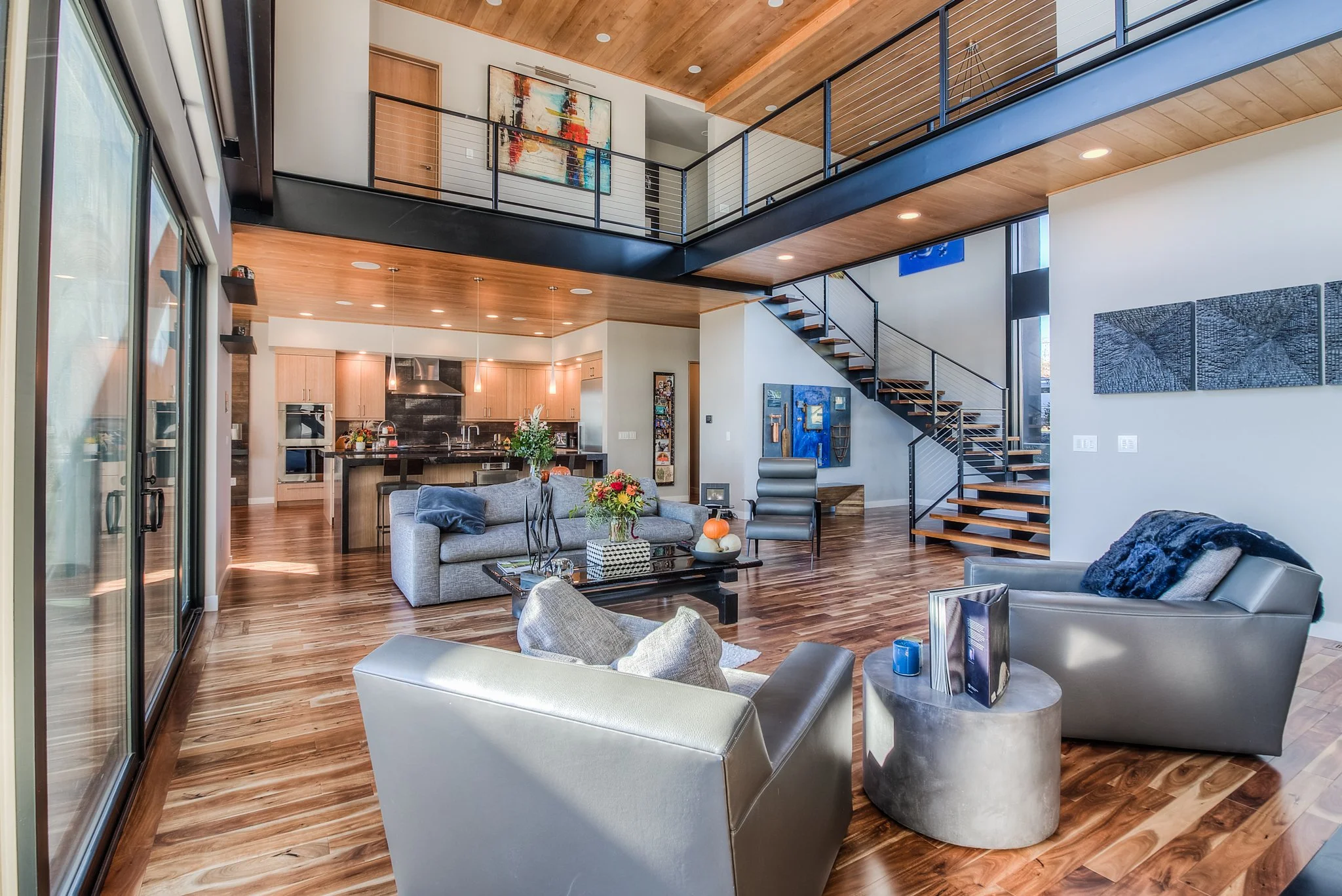 Modern open-concept living room with wooden floors and high ceiling, featuring a kitchen with wooden cabinets and black backsplash, a staircase with black railing, and eclectic wall art.