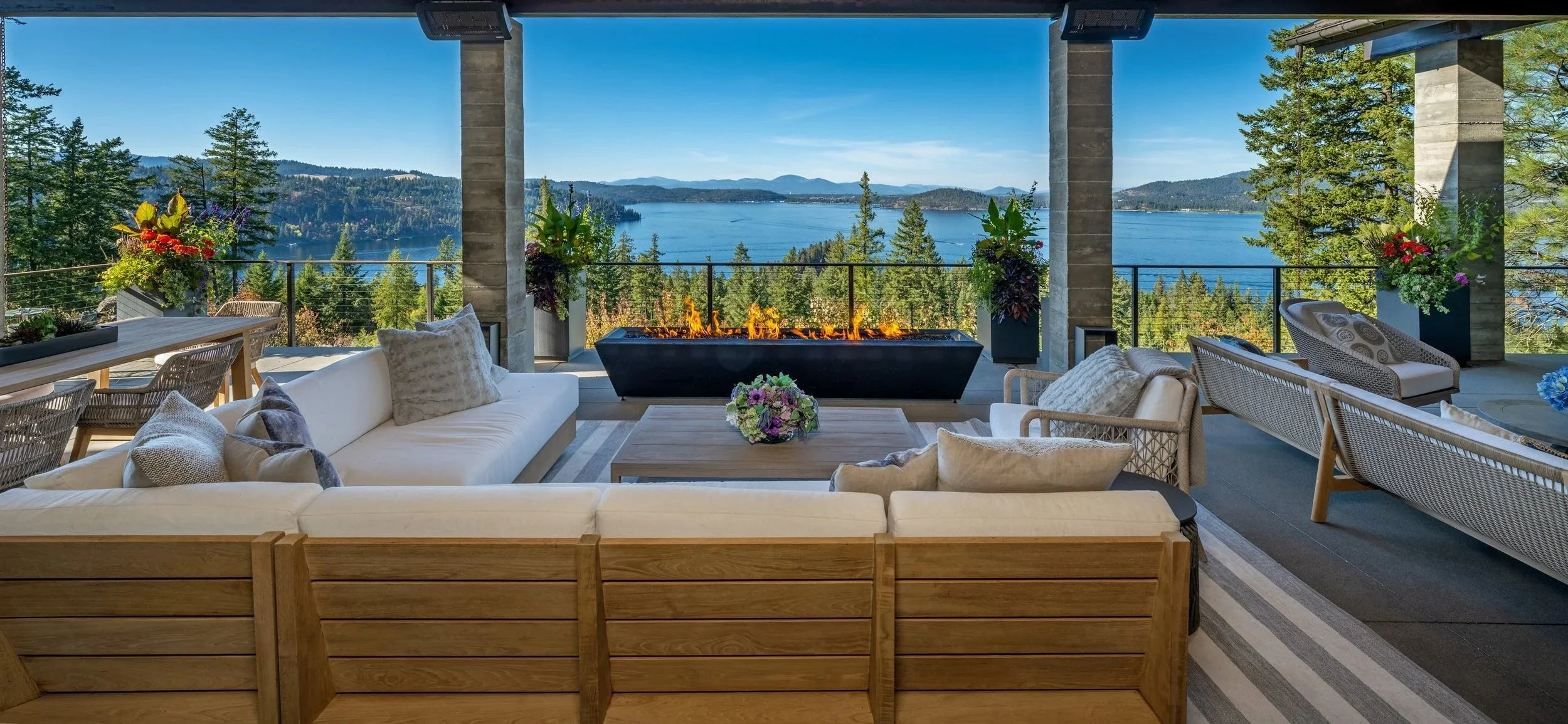 Outdoor patio with white sectional sofa, armchairs, and coffee table, overlooking a lake and mountain range with trees, decorated with colorful flowers and a long orange flames fire pit in the center.