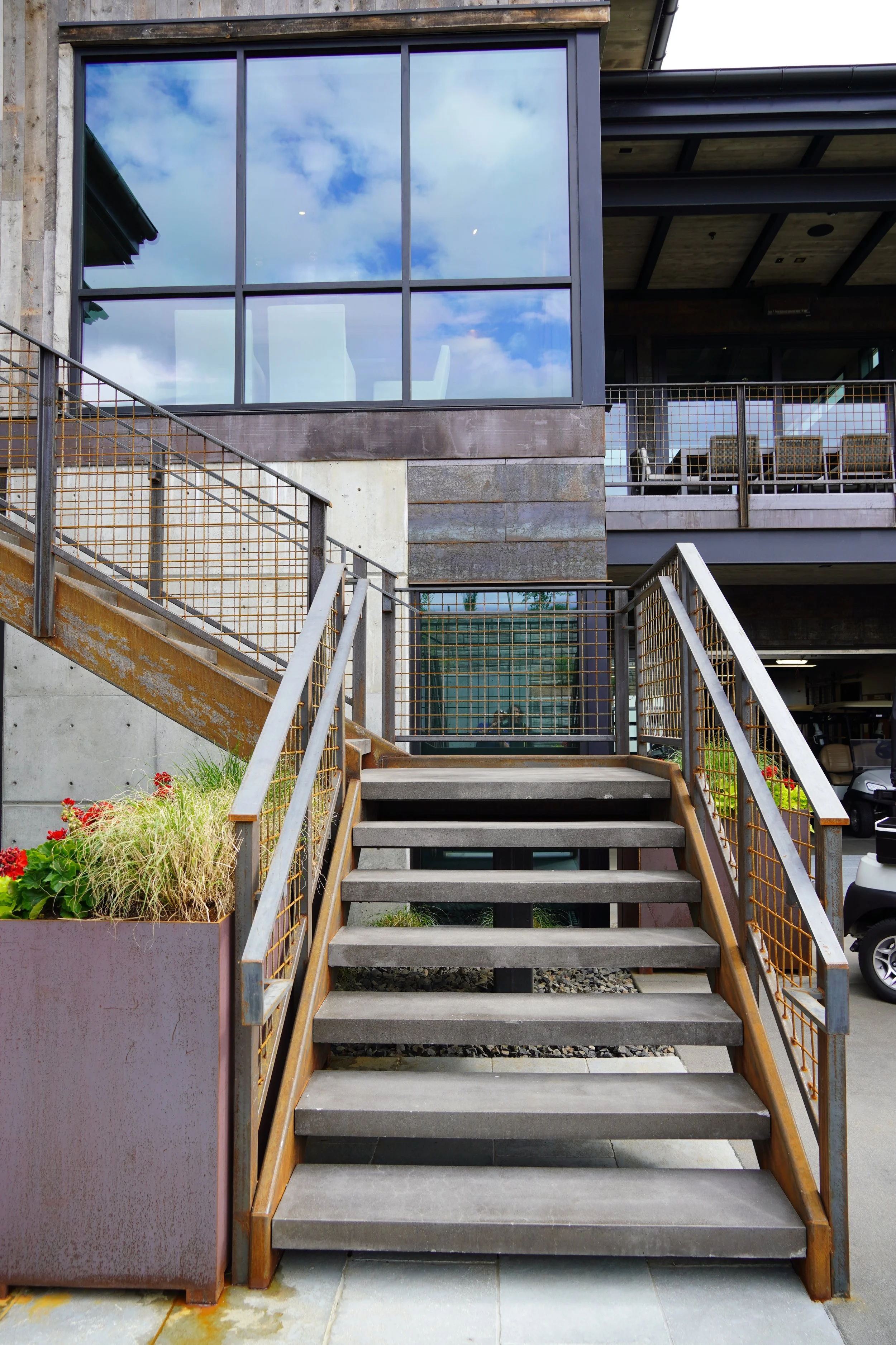 Outdoor staircase with metal railings leading to a modern building with large glass windows reflecting blue sky and clouds.