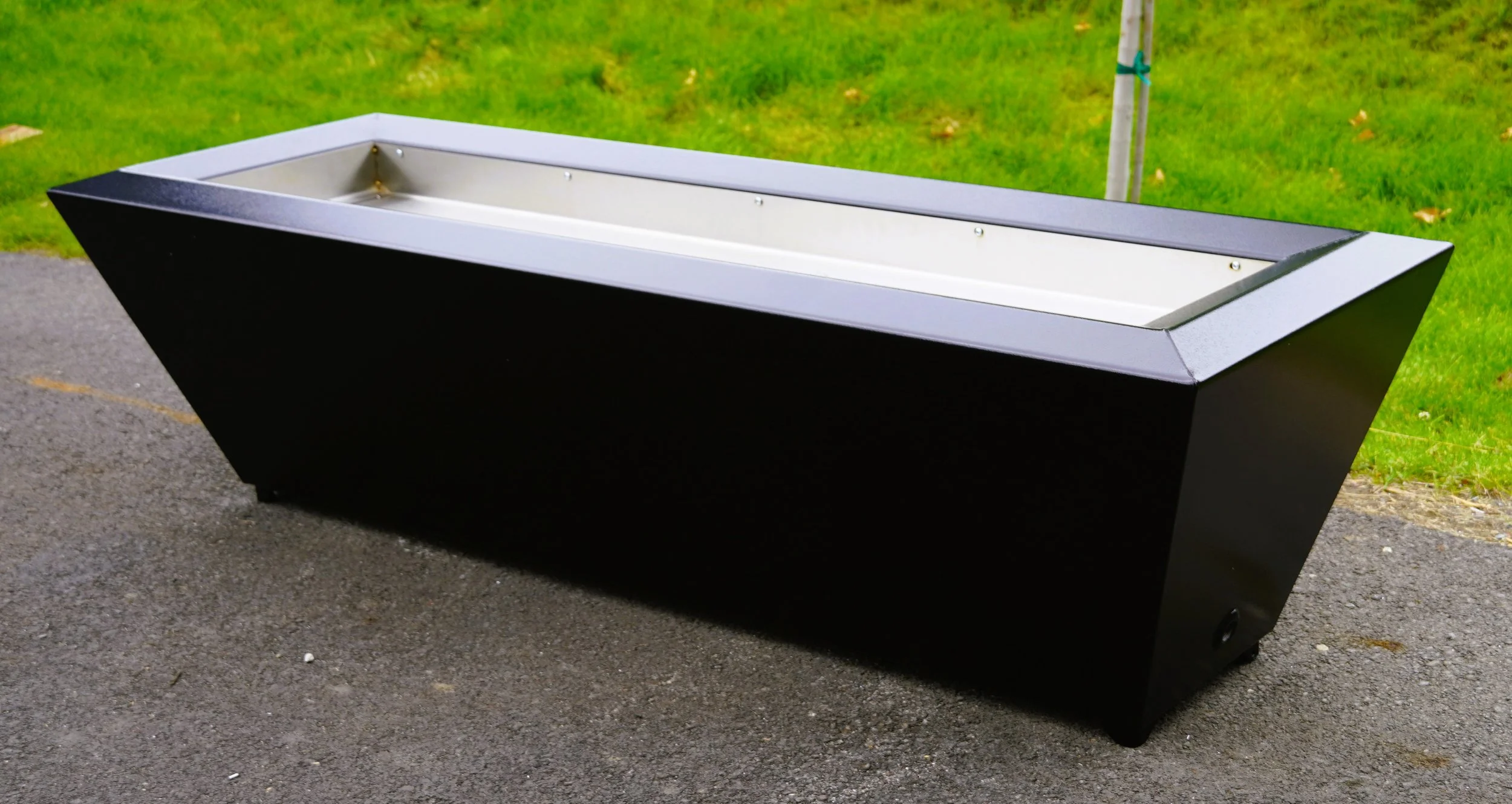 Large black rectangular planter box with a metallic interior placed outdoors on asphalt, with green grass and a tree in the background.