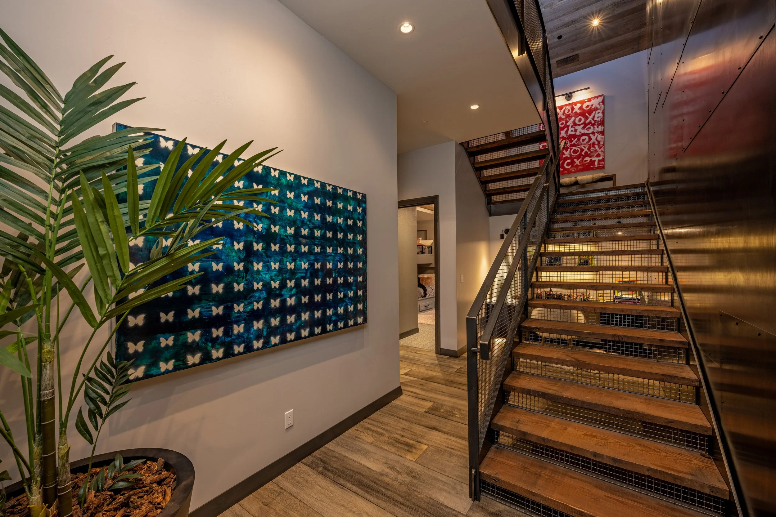 Interior of a modern home with a wooden staircase, a large butterfly artwork on the wall, a potted plant, and a doorway leading to another room.
