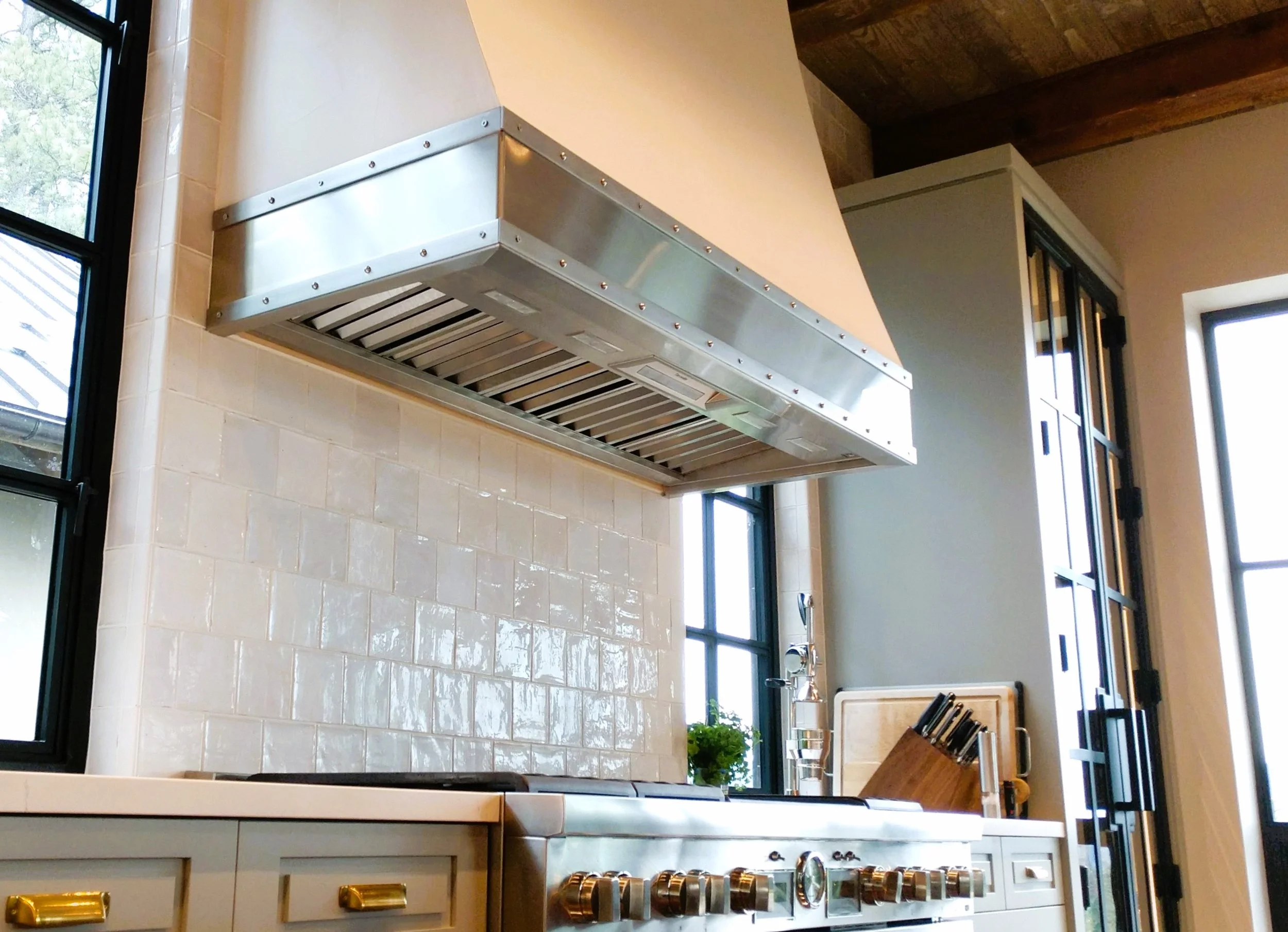 Modern kitchen stove with a stainless steel range hood, white tile backsplash, black window frames, and a potted plant on the counter.
