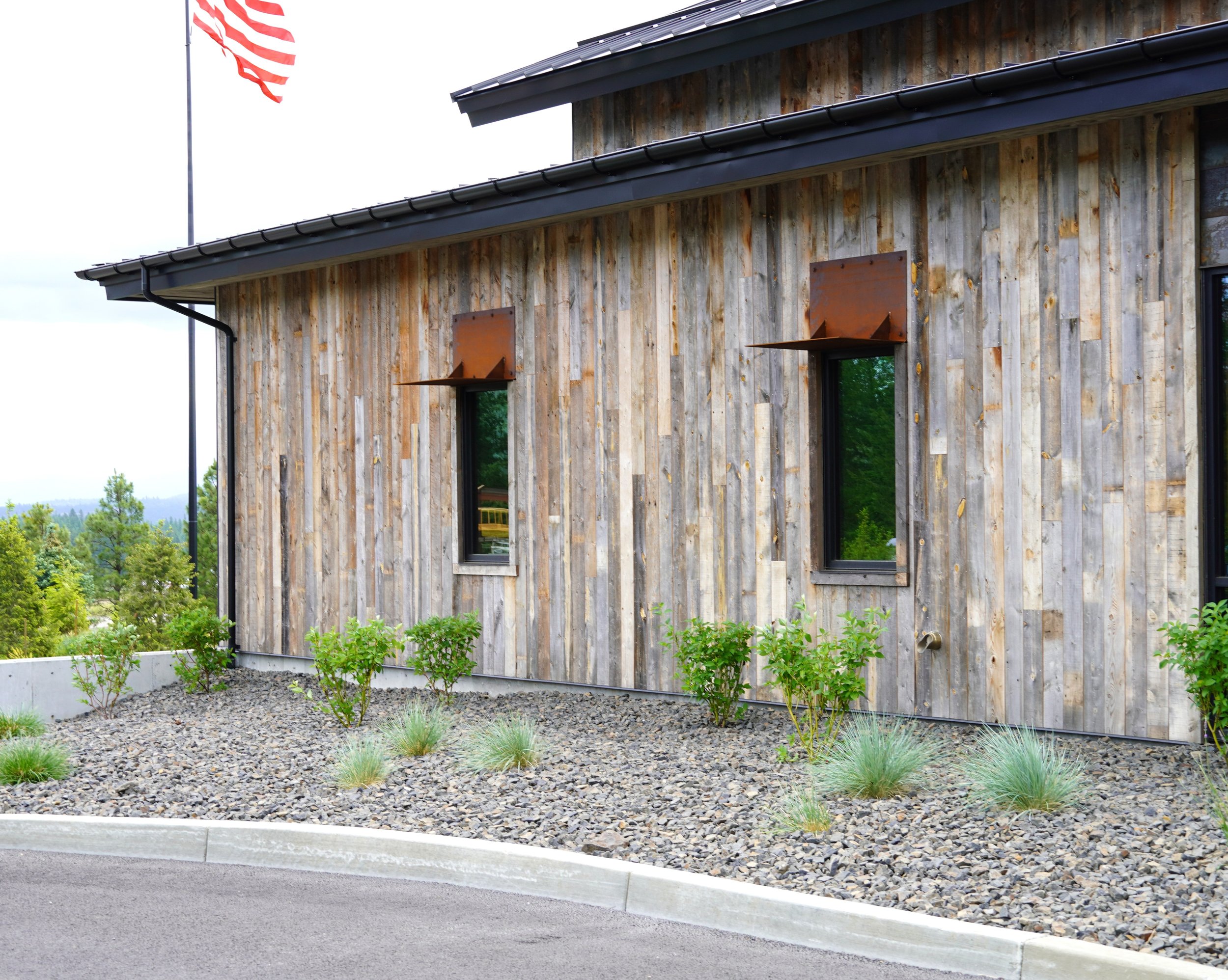 Rustic wooden building with two narrow windows, metal awnings, a sloped metal roof, and surrounded by a landscaped area with small shrubs and grasses.
