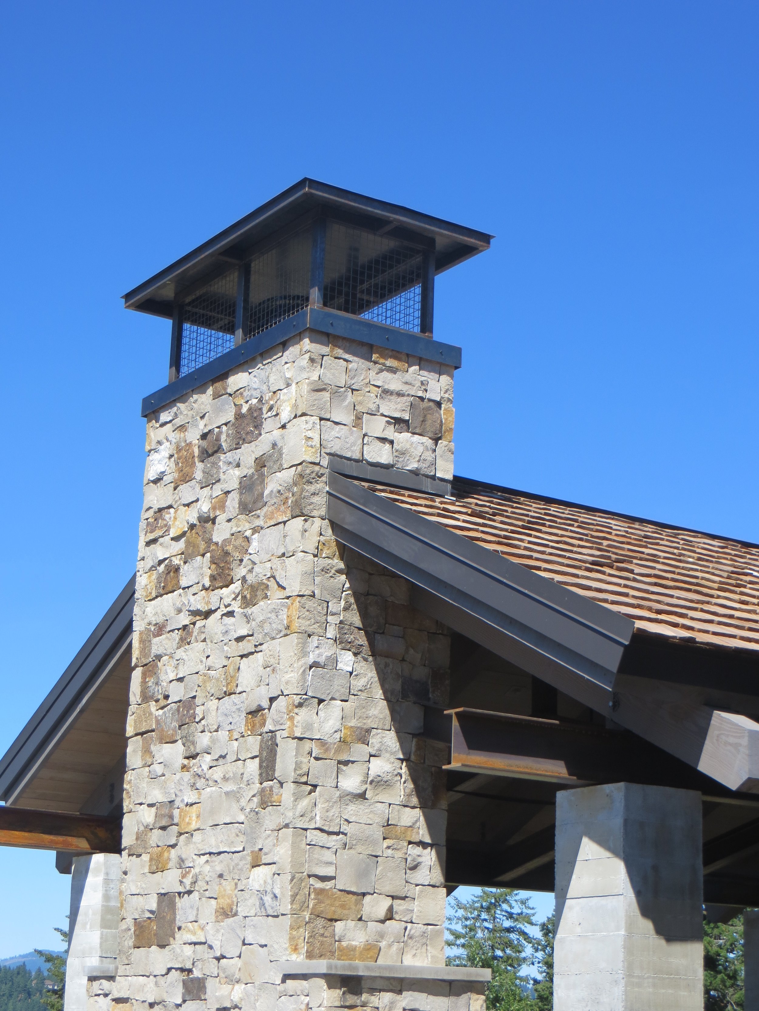 Close-up of a stone chimney with a metal cap and an outdoor shelter roof, with a clear blue sky in the background.