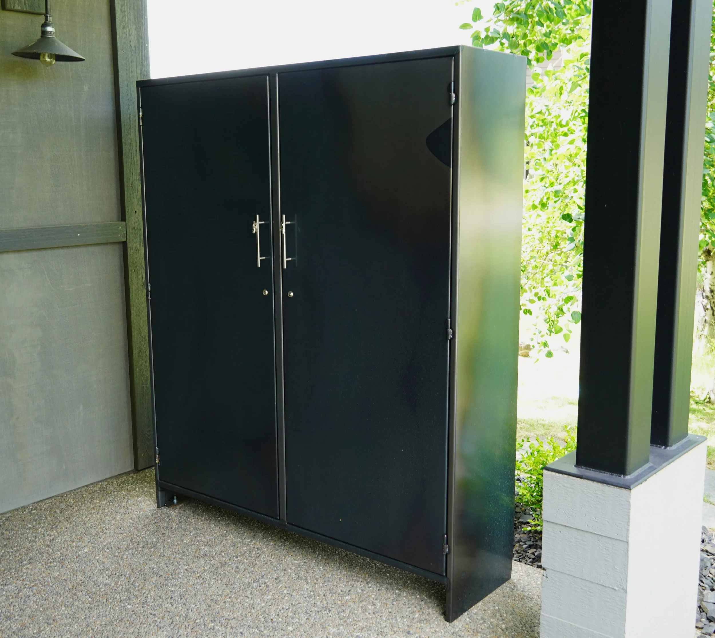 Black metal outdoor storage cabinet on a porch with greenery in the background.