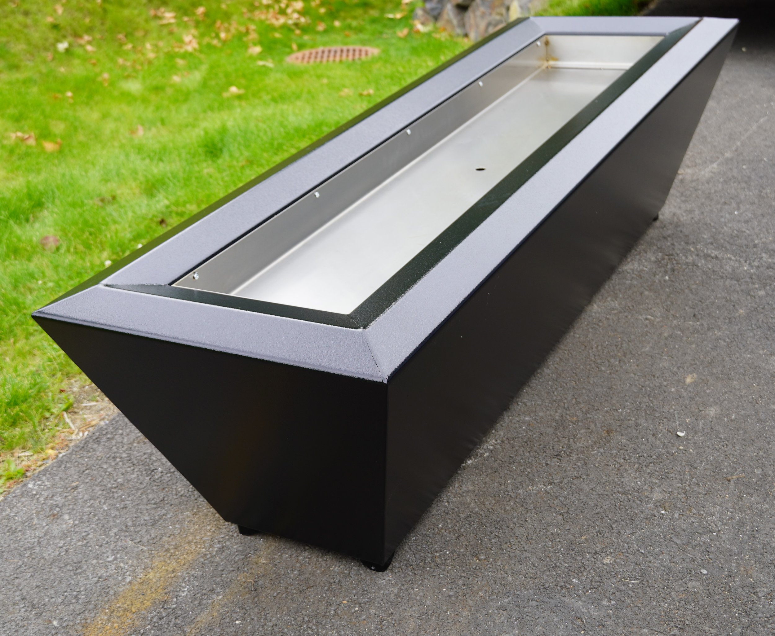 A black rectangular outdoor planter with a glass top, situated on a paved surface near a grassy area.