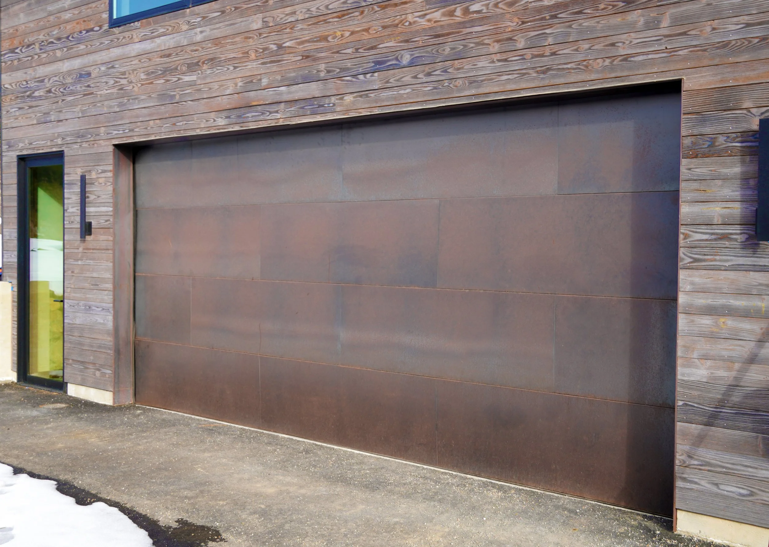 Modern building with a large, rust-colored metal garage door and wood siding. A glass window with a black frame and a handle is visible on the left side of the door, with snow on the ground.