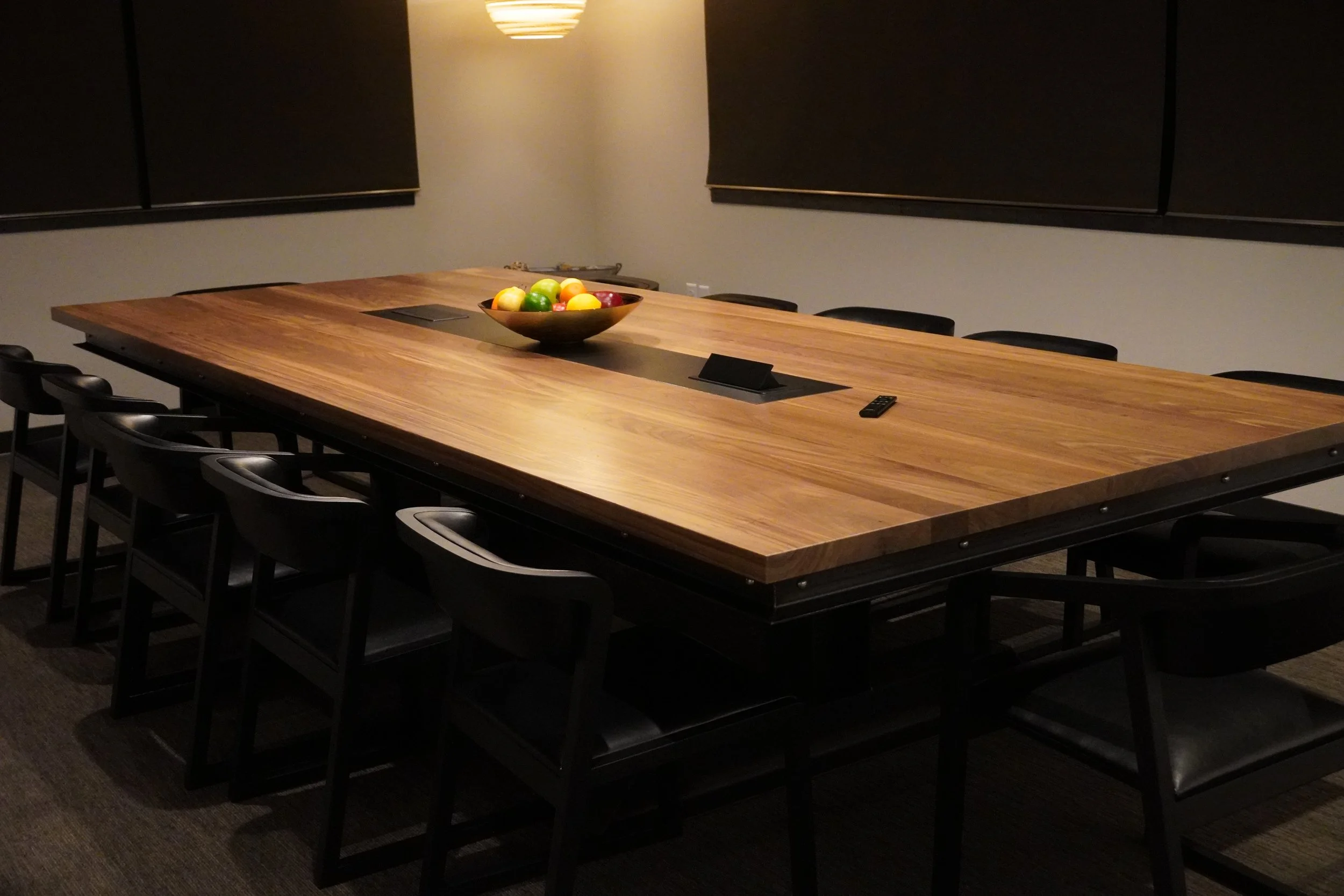 A large wooden conference table with nine black chairs around it in a dimly lit room. There is a bowl of assorted fruits in the center of the table and a remote control on the table. The walls are plain and the windows have black blinds.