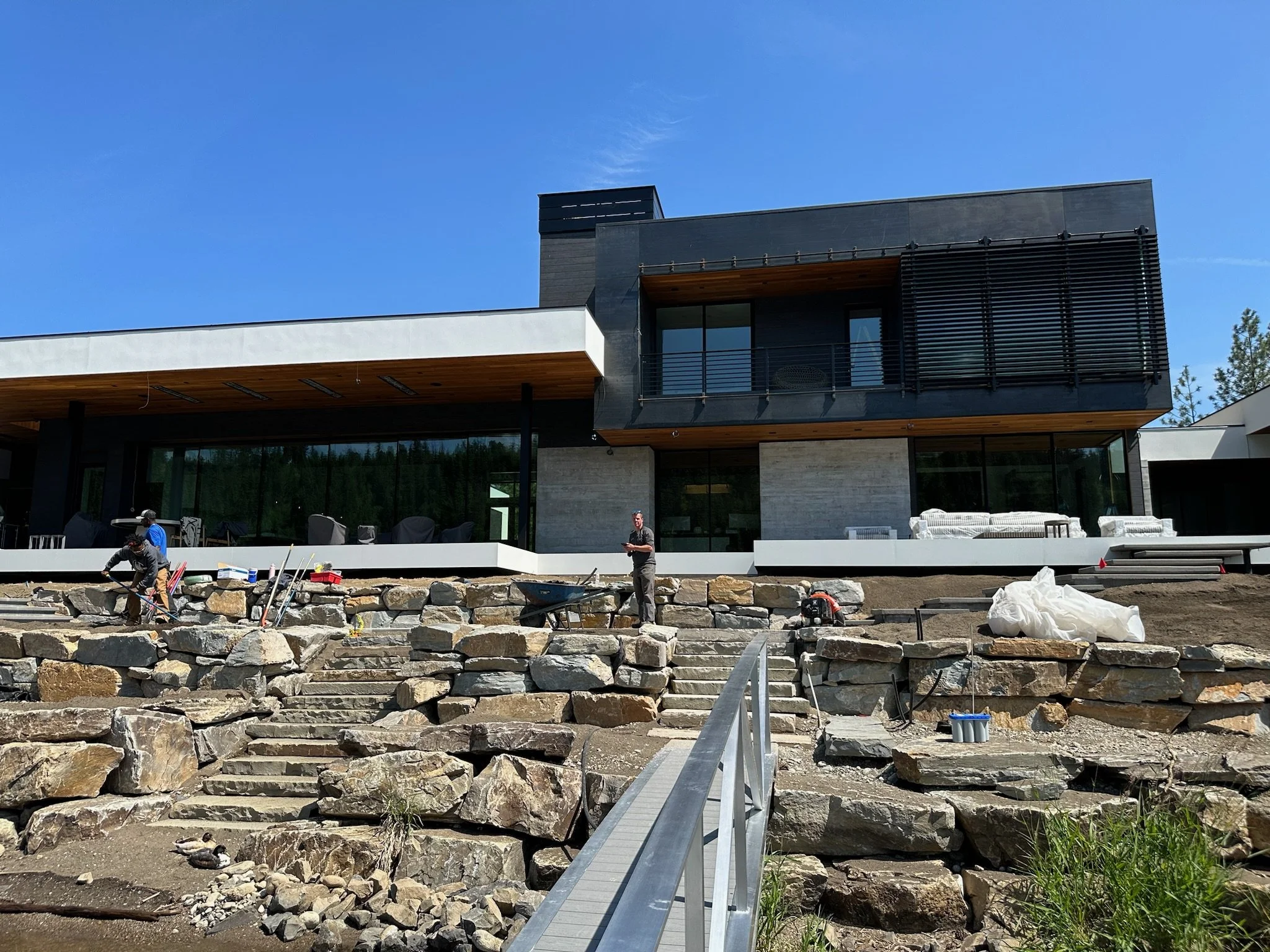 Construction workers building a stone pathway in front of a modern house with large windows and a balcony under a clear blue sky.