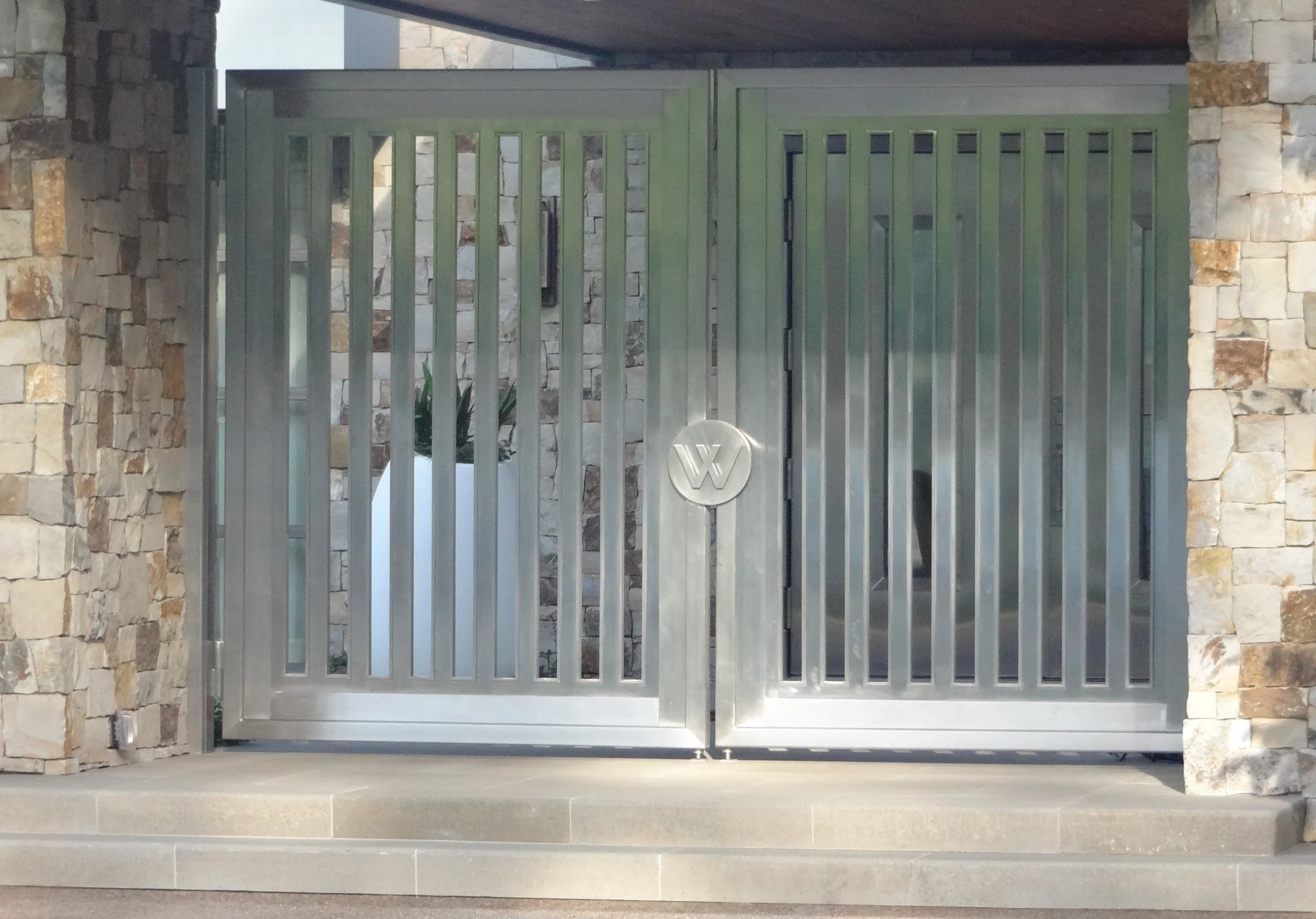 Metal gate with vertical bars and a circular emblem in the center, set between stone walls with a plant visible through the gate.