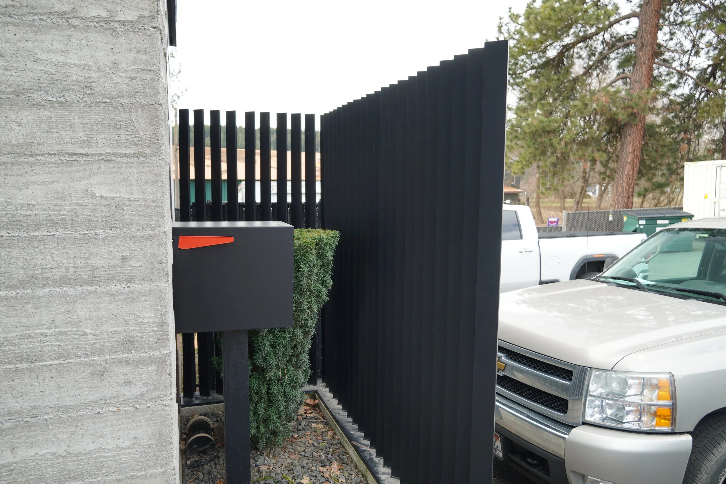 A concrete wall on the left with a black metal gate and a bush underneath it. Behind the gate, a white pickup truck and a silver truck are parked outside in a lot, with trees and other vehicles visible in the background.