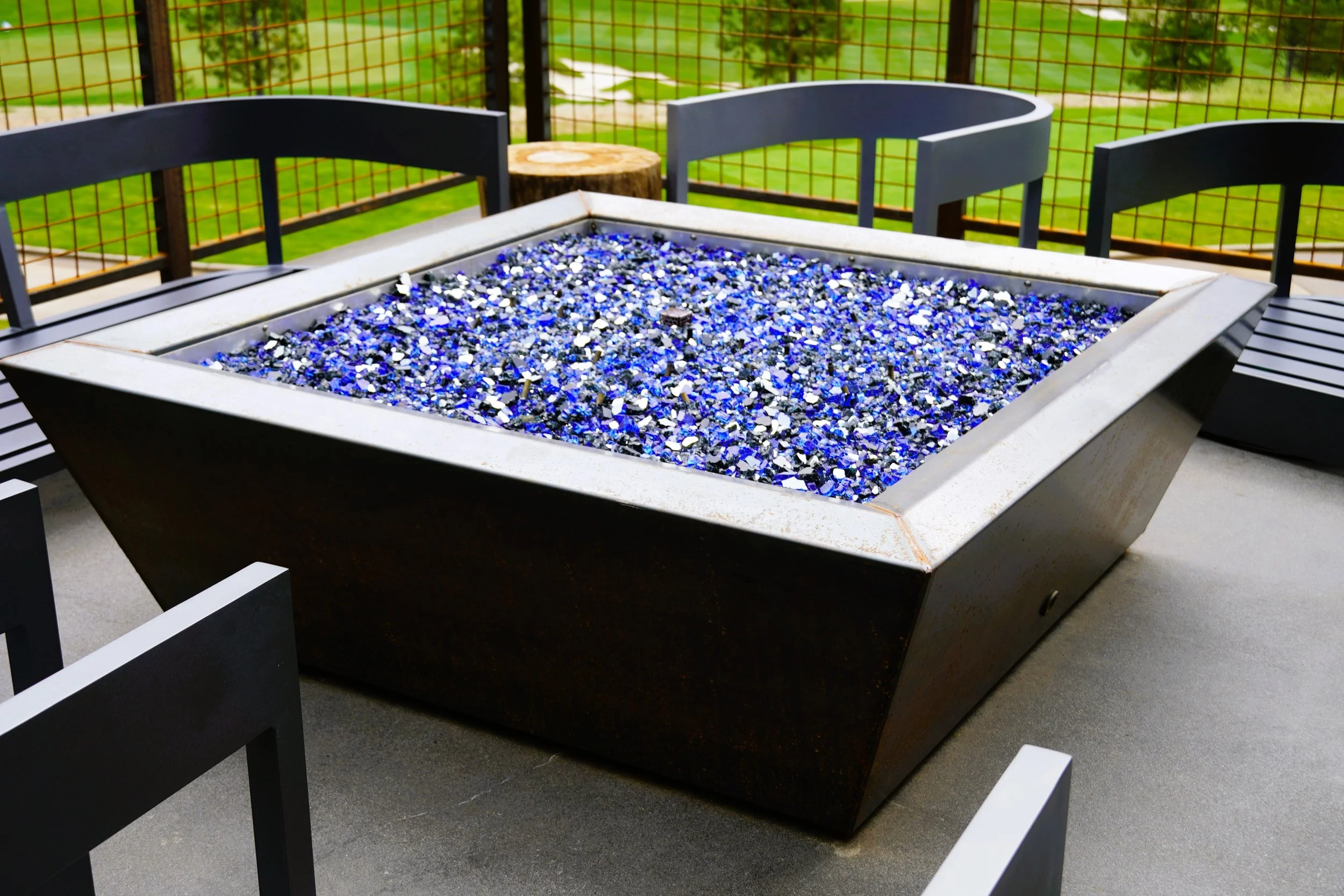 Outdoor patio table with a fire pit filled with blue and black glass pieces, surrounded by black chairs, with a grassy yard and fence in the background.