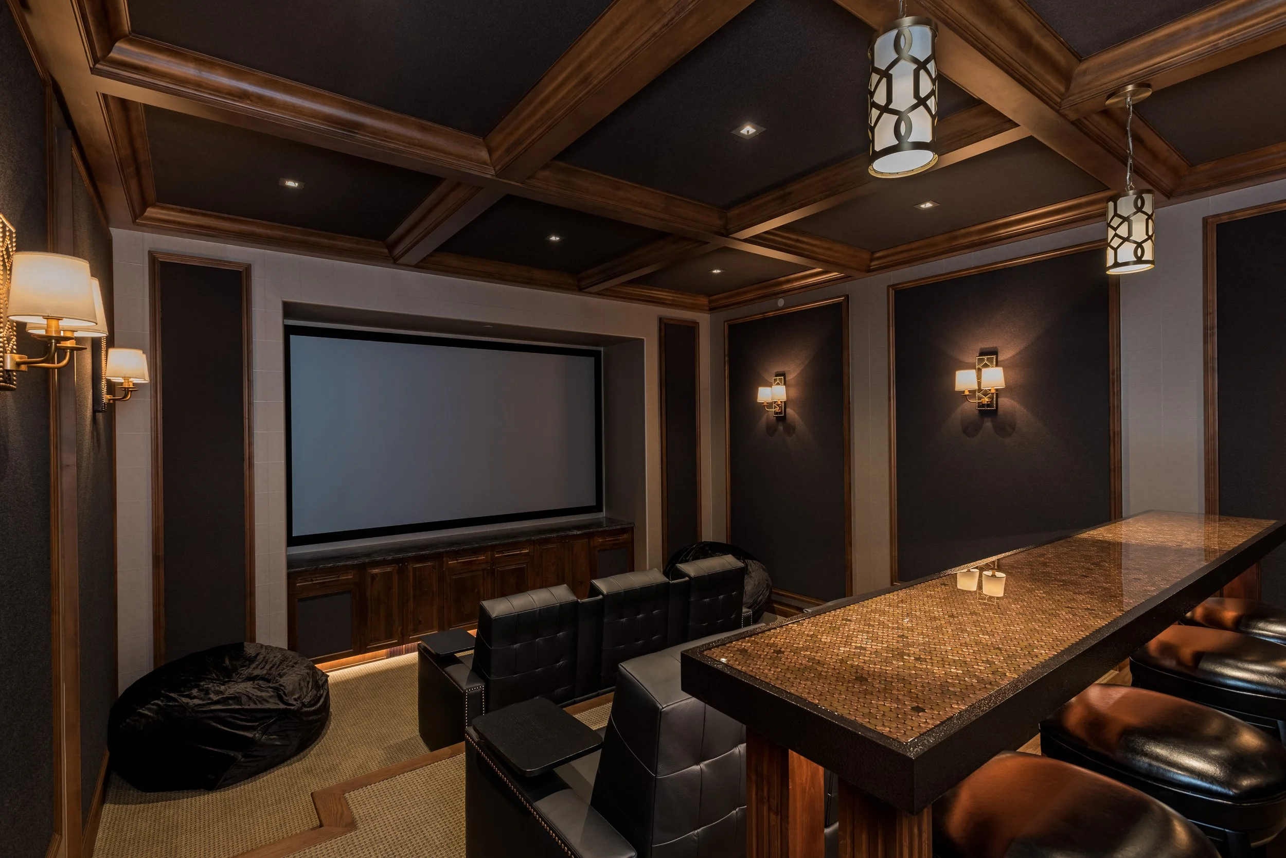 A home theater room with a large screen, black leather chairs, a bar counter with bar stools, dim wall sconces, and a coffered ceiling with modern light fixtures.