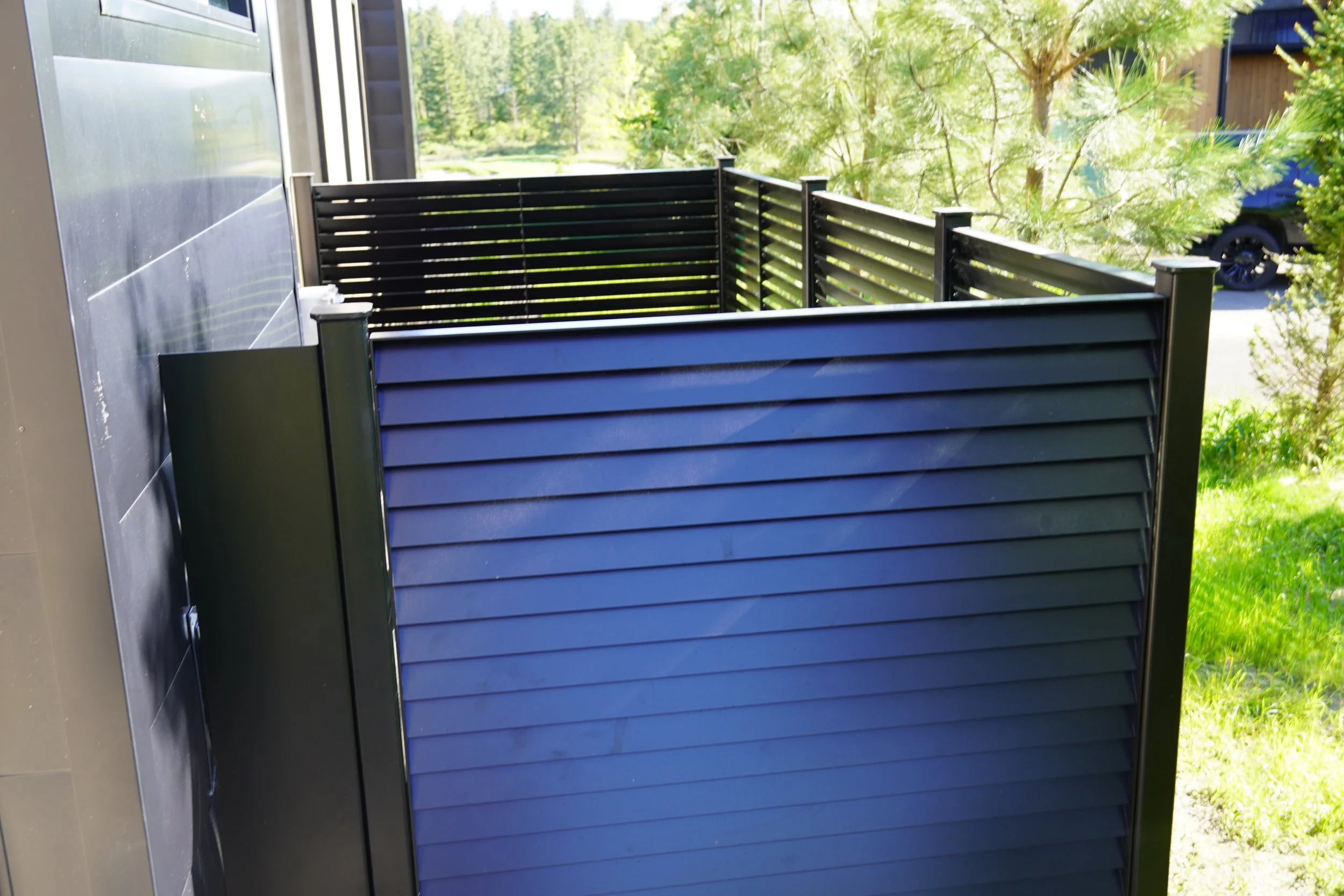 A small outdoor balcony or patio with horizontal black slat railing, attached to a building with dark gray siding, overlooking a green landscape with trees and bushes.
