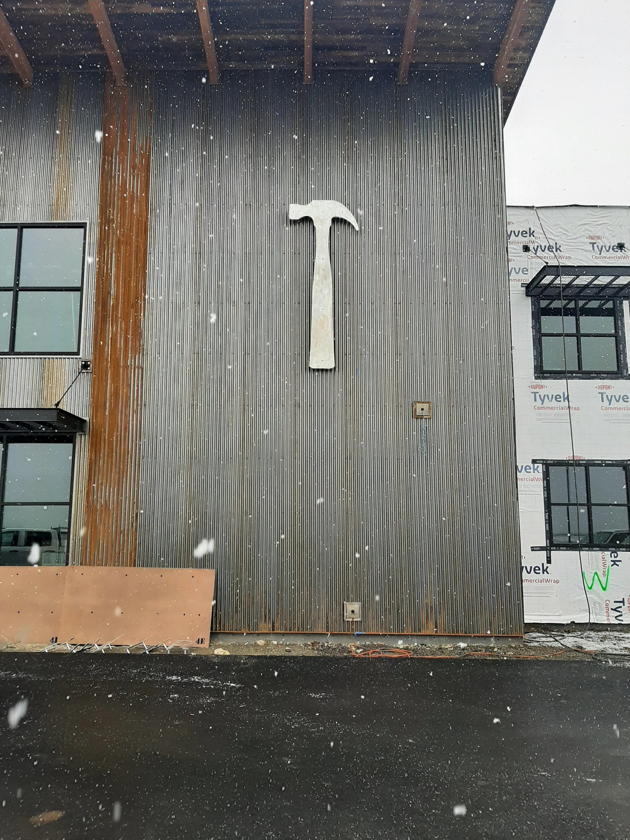 Front exterior of a building under construction with a large hammer logo mounted on a corrugated metal wall, surrounded by snow.