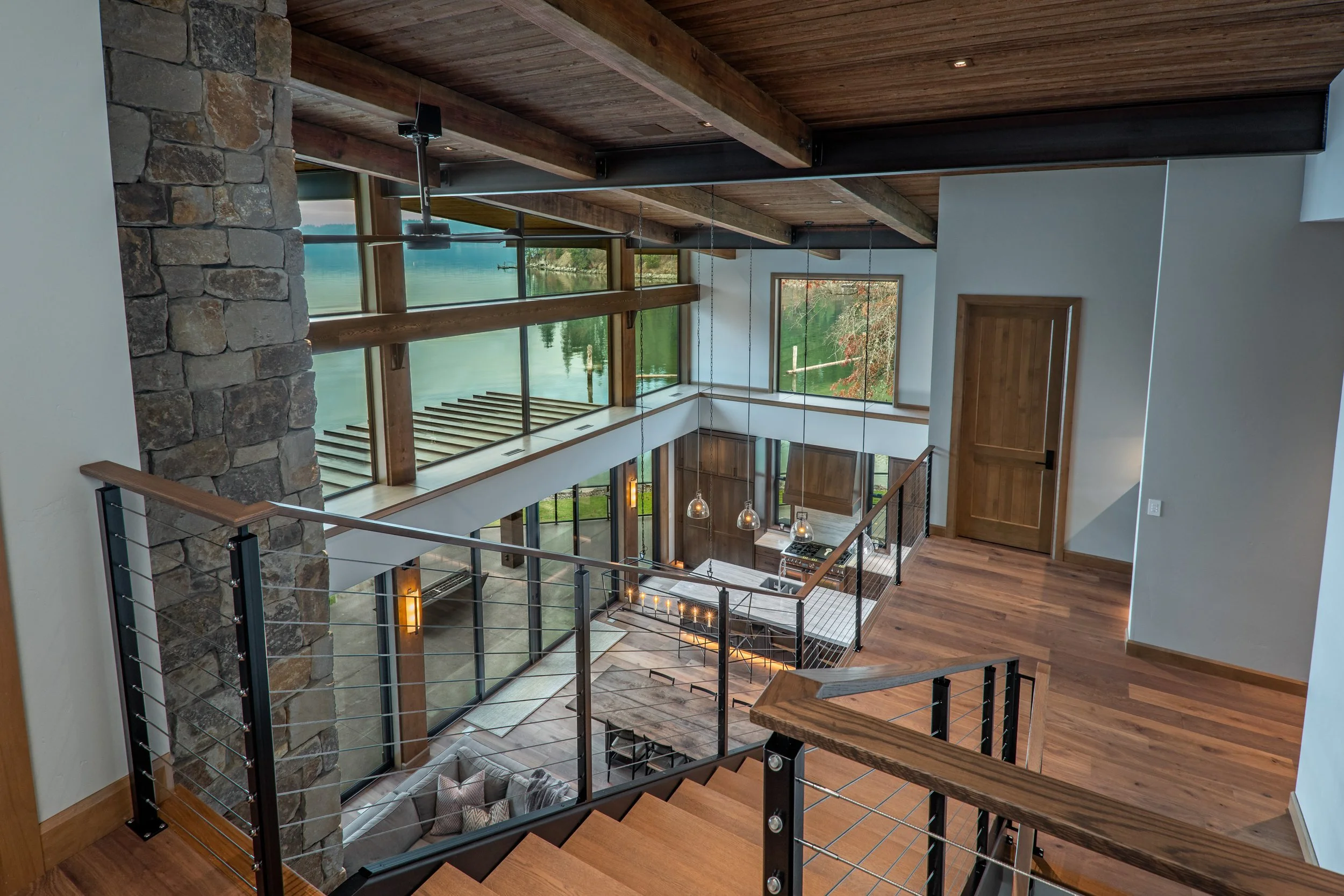 A modern open-concept home interior featuring a staircase with black metal railing, wooden floors, large windows overlooking a lake, and a stone accent wall.