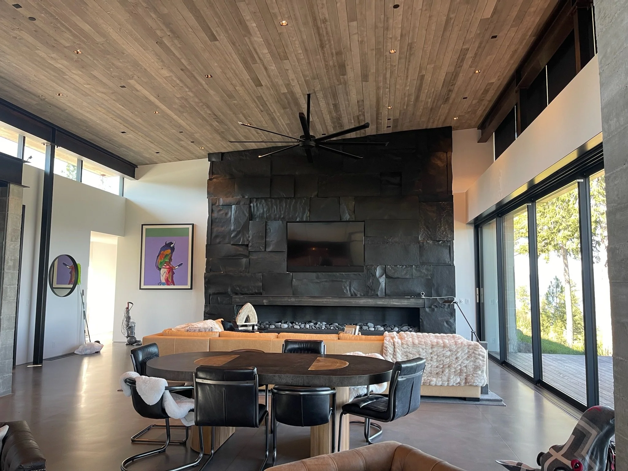 Modern living room with black stone fireplace, wooden ceiling, large windows, round dining table with black chairs, and art on the wall.