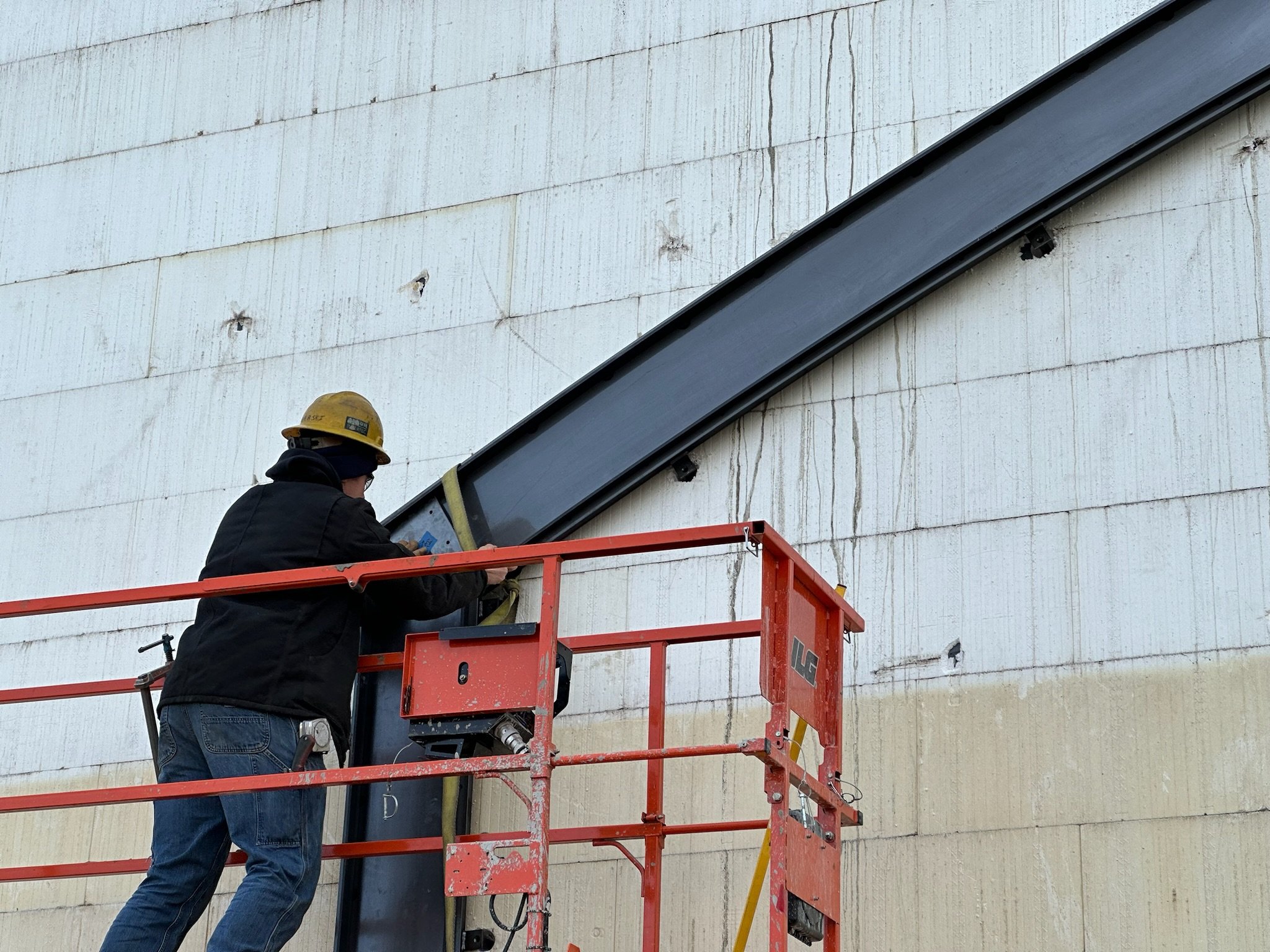 Worker wearing a yellow safety helmet and black jacket standing on a red lift platform, working on a large structure attached to the side of a building.