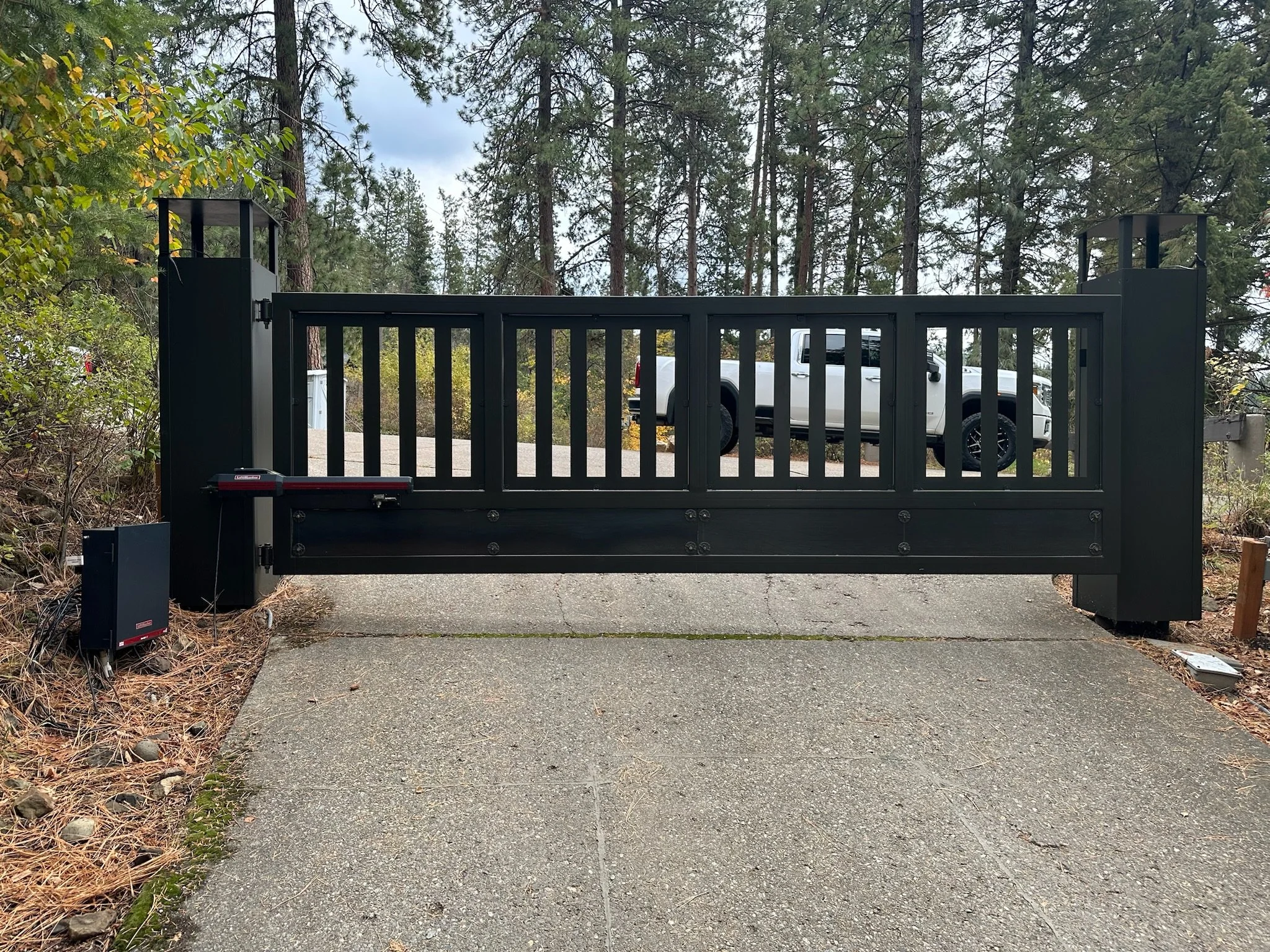 A black automated sliding gate on a concrete driveway with a white pickup truck behind it in a forested area.