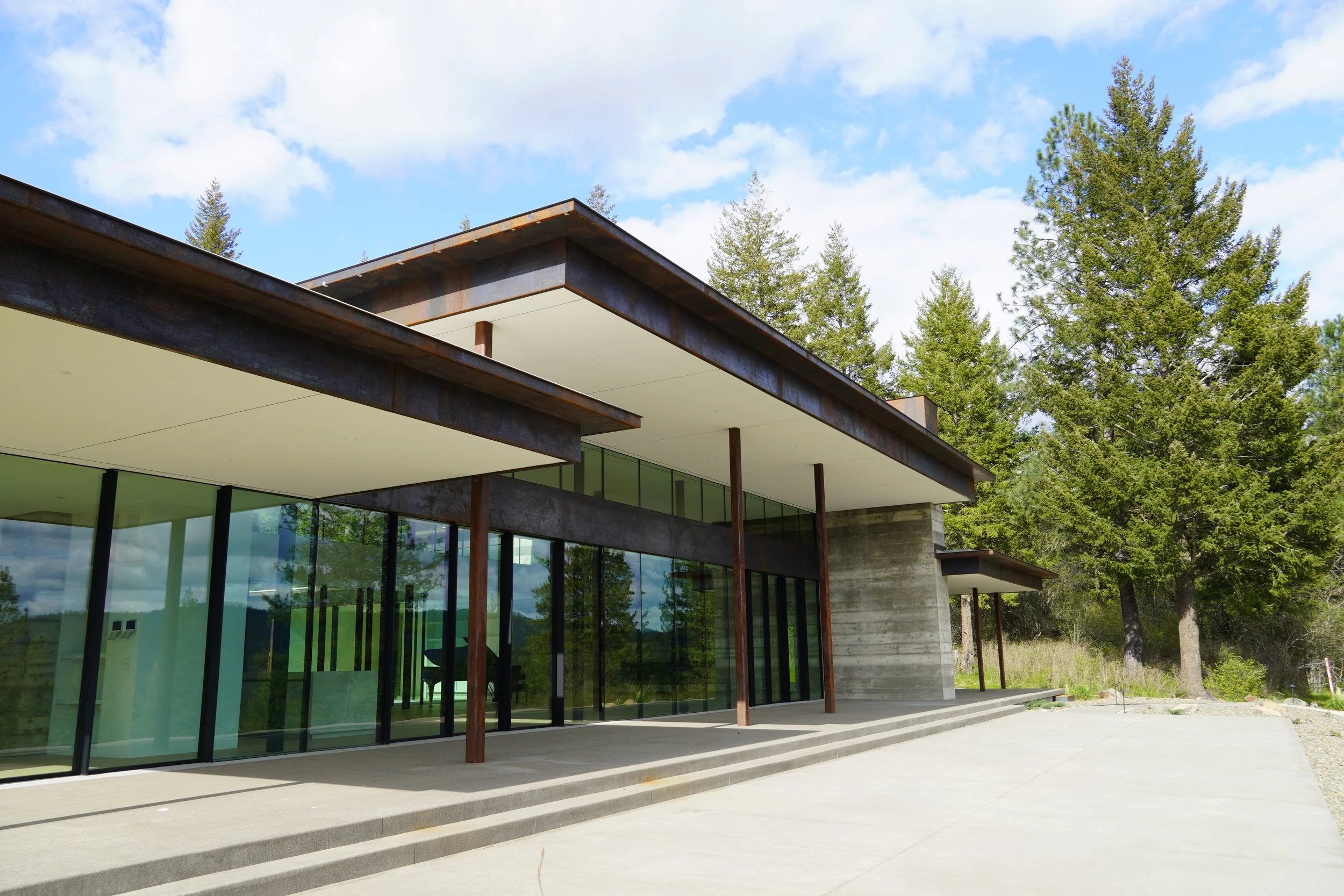 Modern building with large glass windows and a cantilevered roof, surrounded by green trees and a cloudy sky.