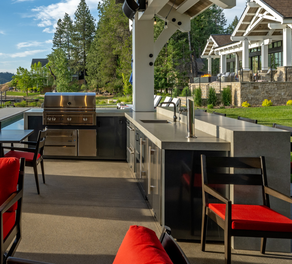 Outdoor kitchen with a grill, countertop, and seating area, overlooking a green yard with trees and a modern house in the background.