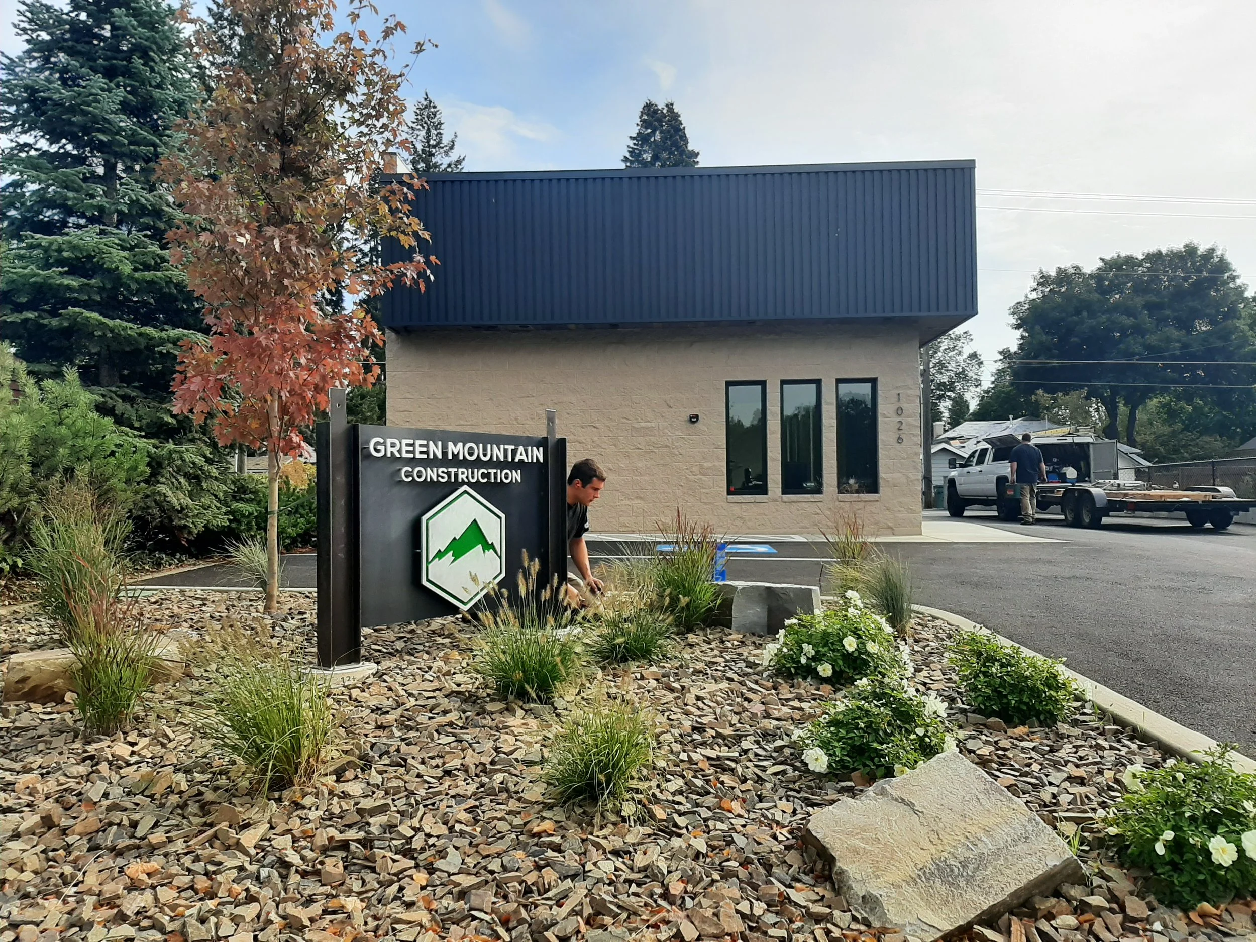 A construction site in front of a building with a sign that reads 'Green Mountain Construction.' There is a young man working near the sign, which is surrounded by rocks and plants. A white pickup truck with a trailer is parked in the background.