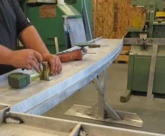 Person working on a curved surfboard in a woodworking shop, with tools and machinery around.