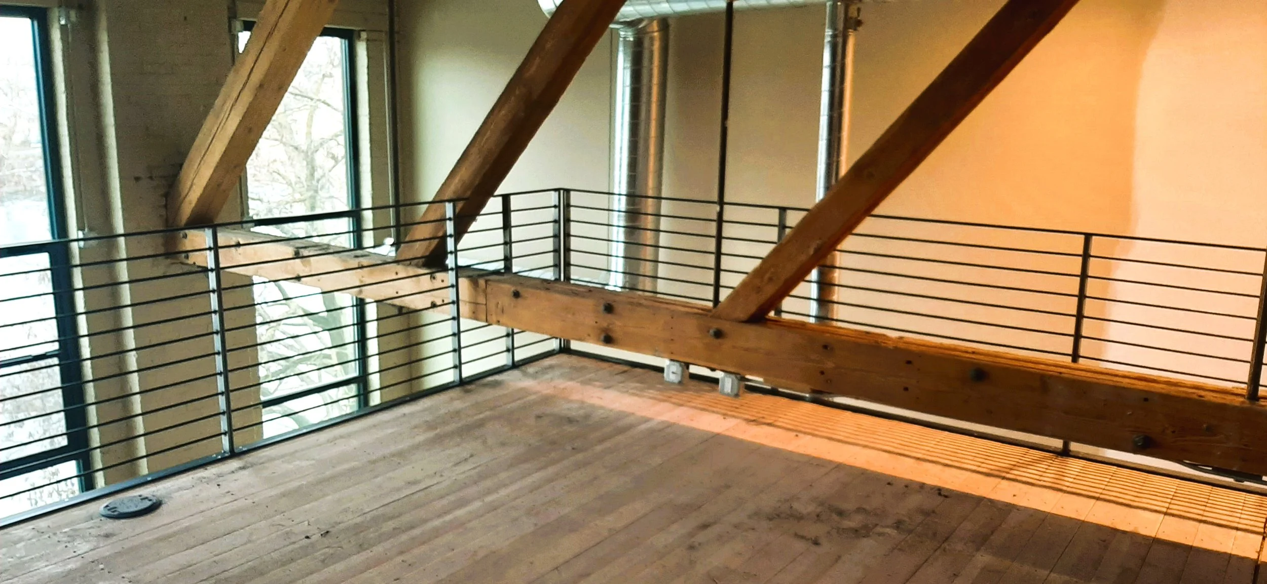 Interior of a room with wooden beams and flooring, large windows showing trees outside, metal pipes, and a black metal railing around the edge of a balcony or loft area.