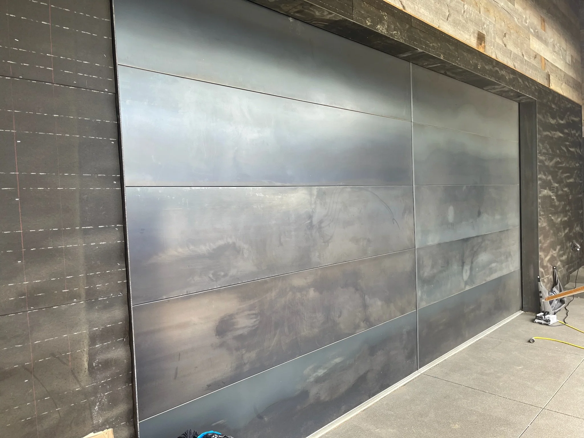 Large metallic sliding door or panel with reflective surface, surrounding wall with textured stone finish, flooring with smooth tiles, and tools or equipment nearby.