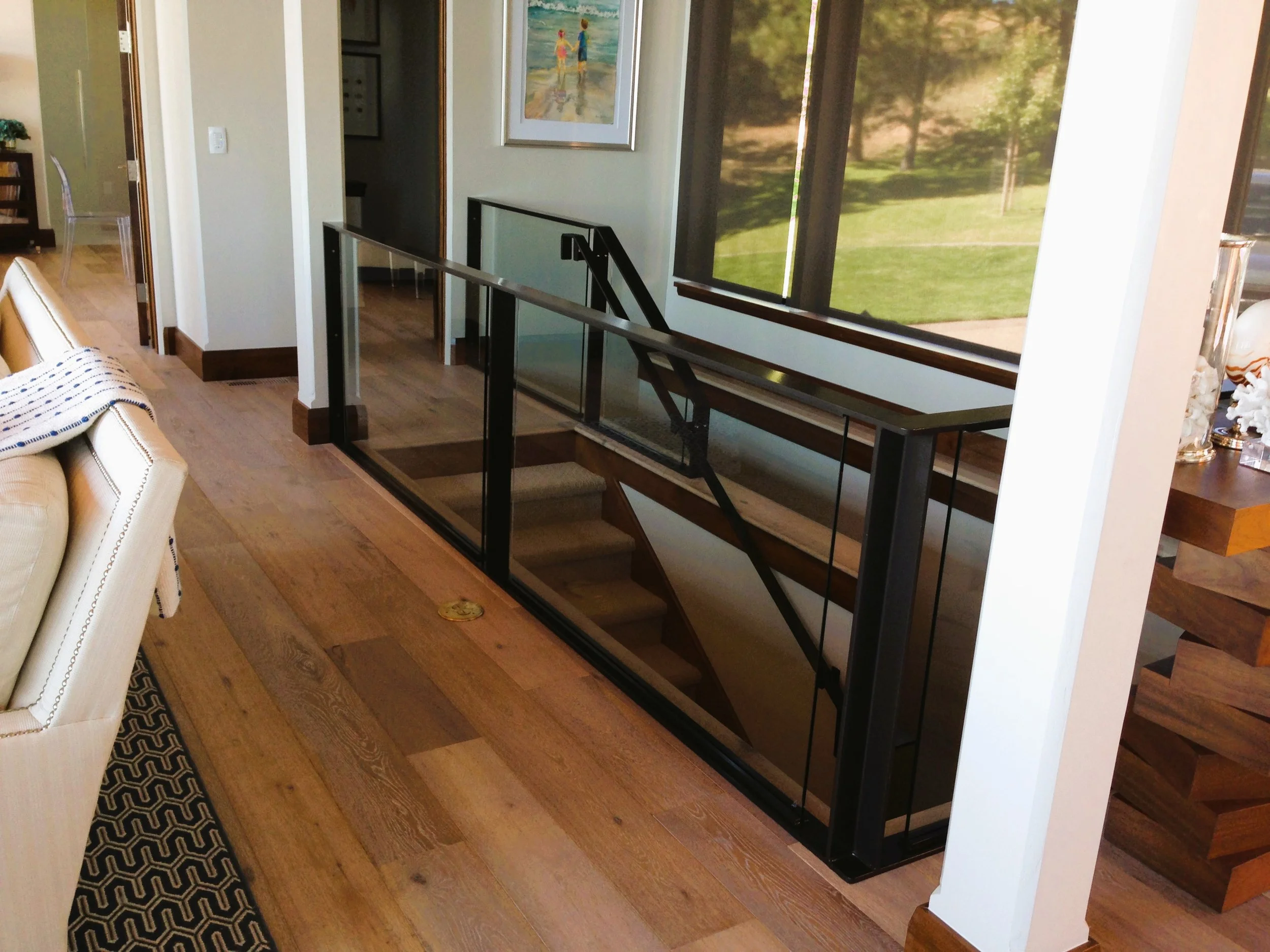 Interior view of a living space with hardwood floors, a glass railing along a staircase, a large window revealing trees outside, a painting on the wall, a cream-colored couch partially visible on the left, and a decorative black and cream mat on the 