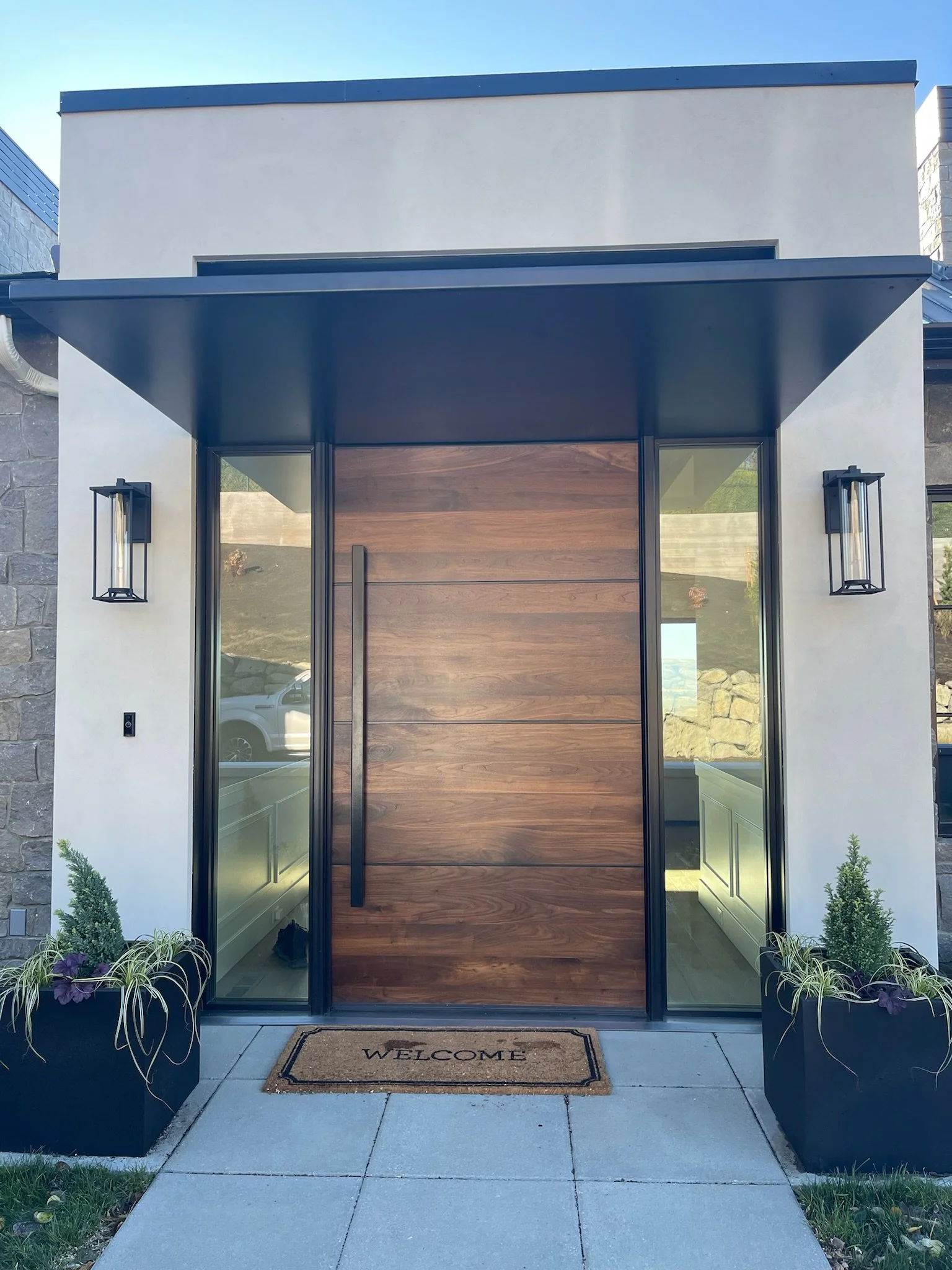 Modern front door with wooden panels, flanked by glass windows, black metal fixtures, potted plants, and a welcome mat.
