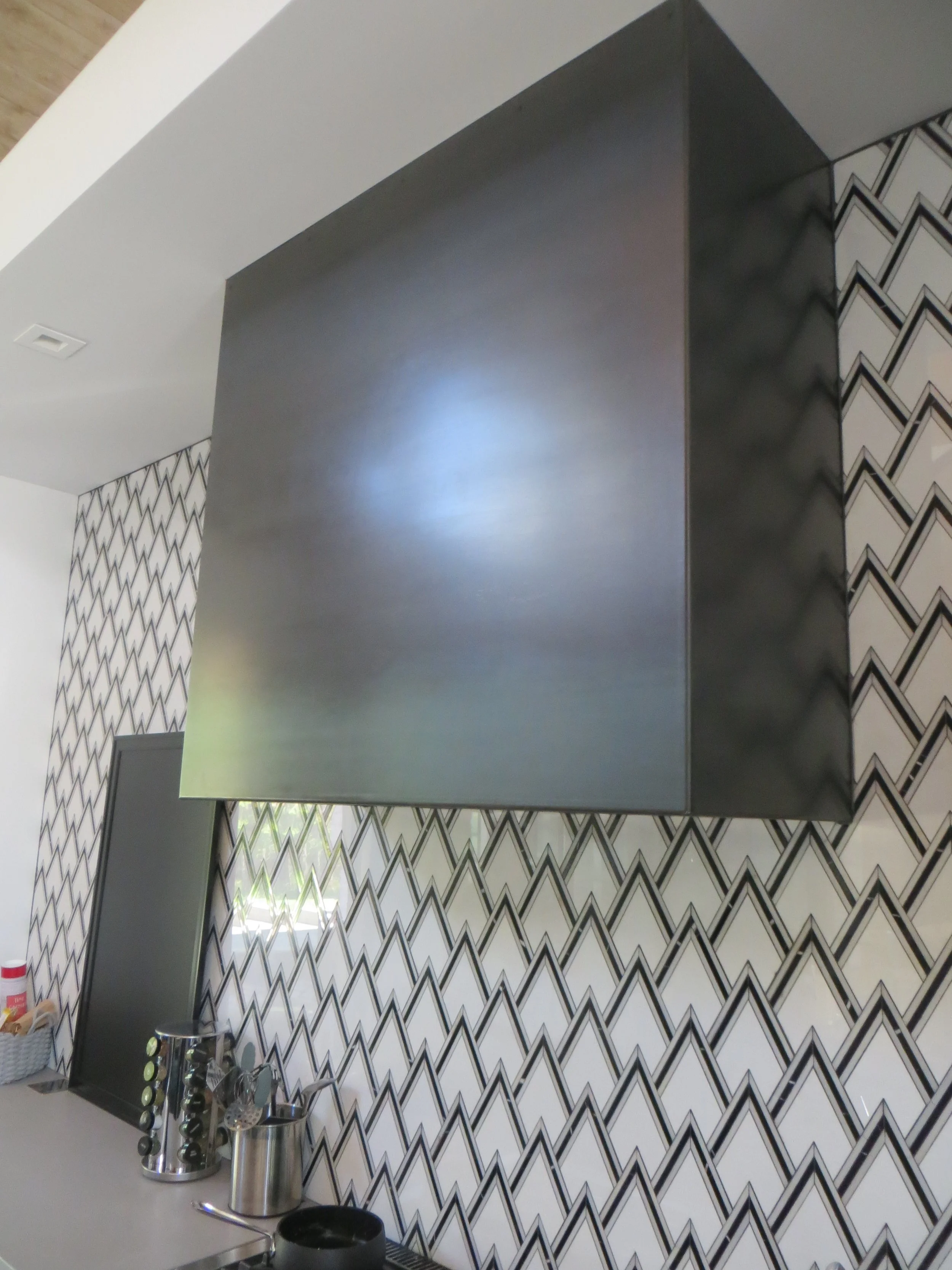 Modern kitchen with a geometric black and white tiled backsplash, stainless steel range hood, and countertop with utensils and containers.