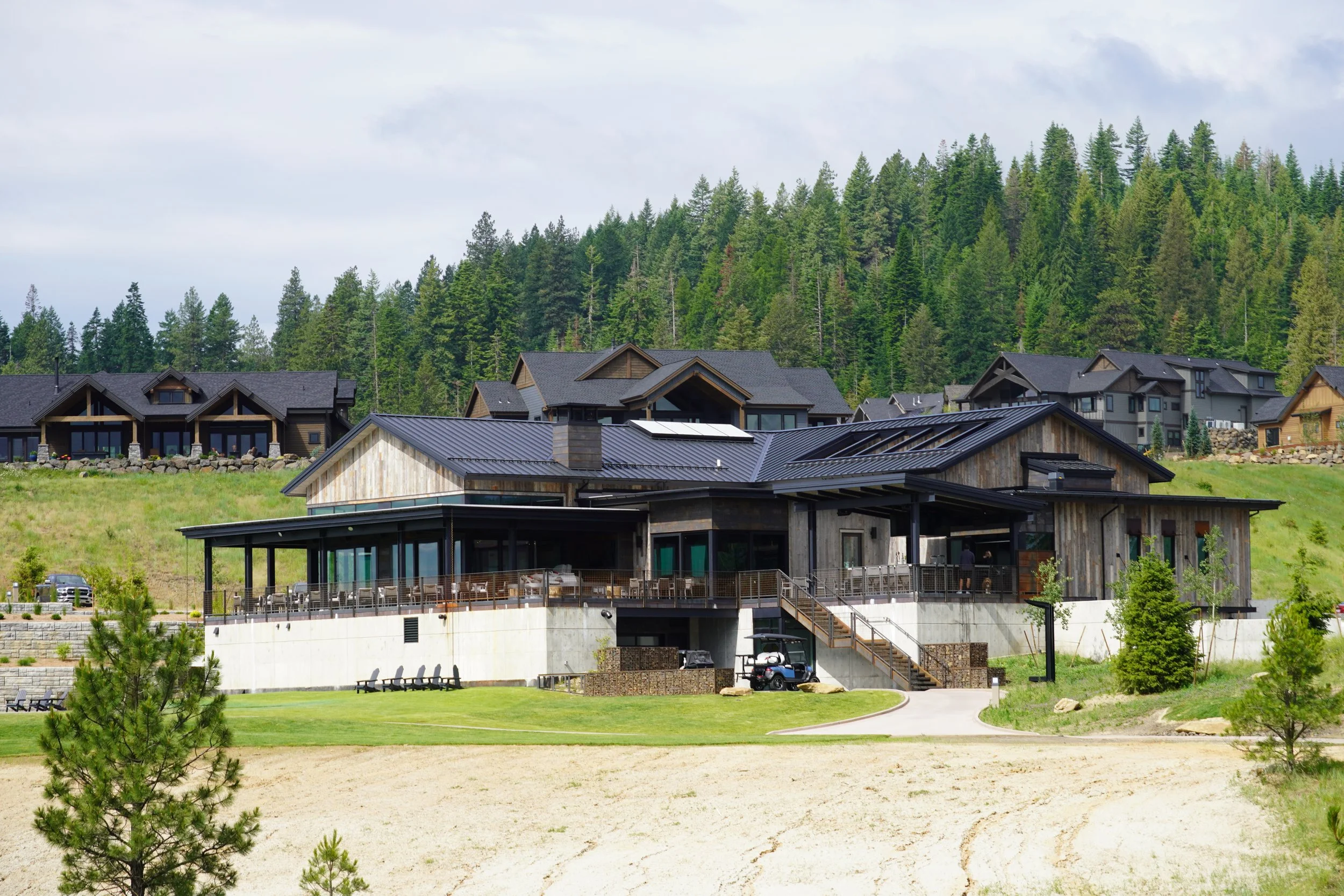 Large modern house with outdoor patio, surrounded by green grass, trees, and neighboring houses on a hillside.