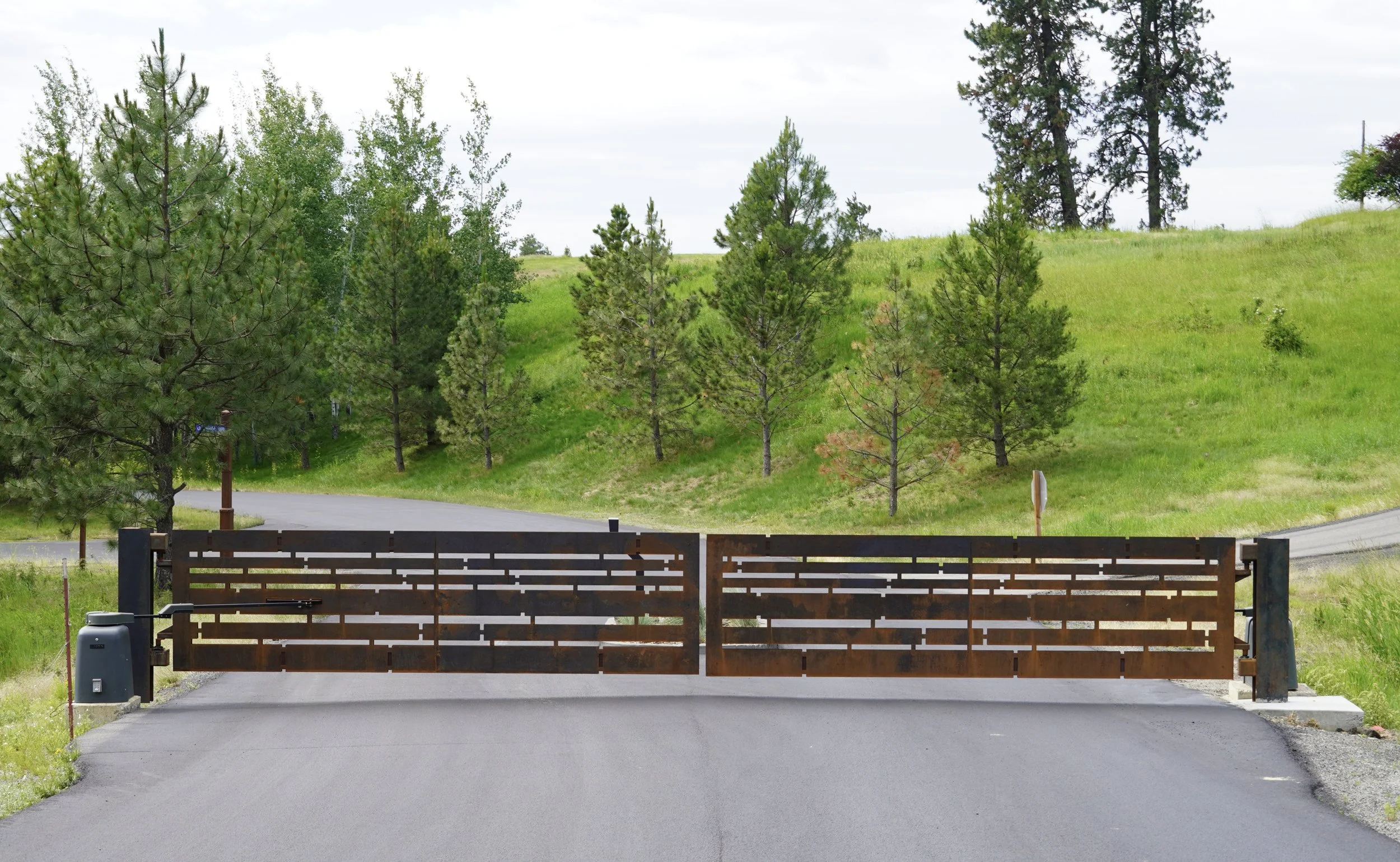 A metal gate blocking a paved road surrounded by green grass and trees with rolling hills in the background.