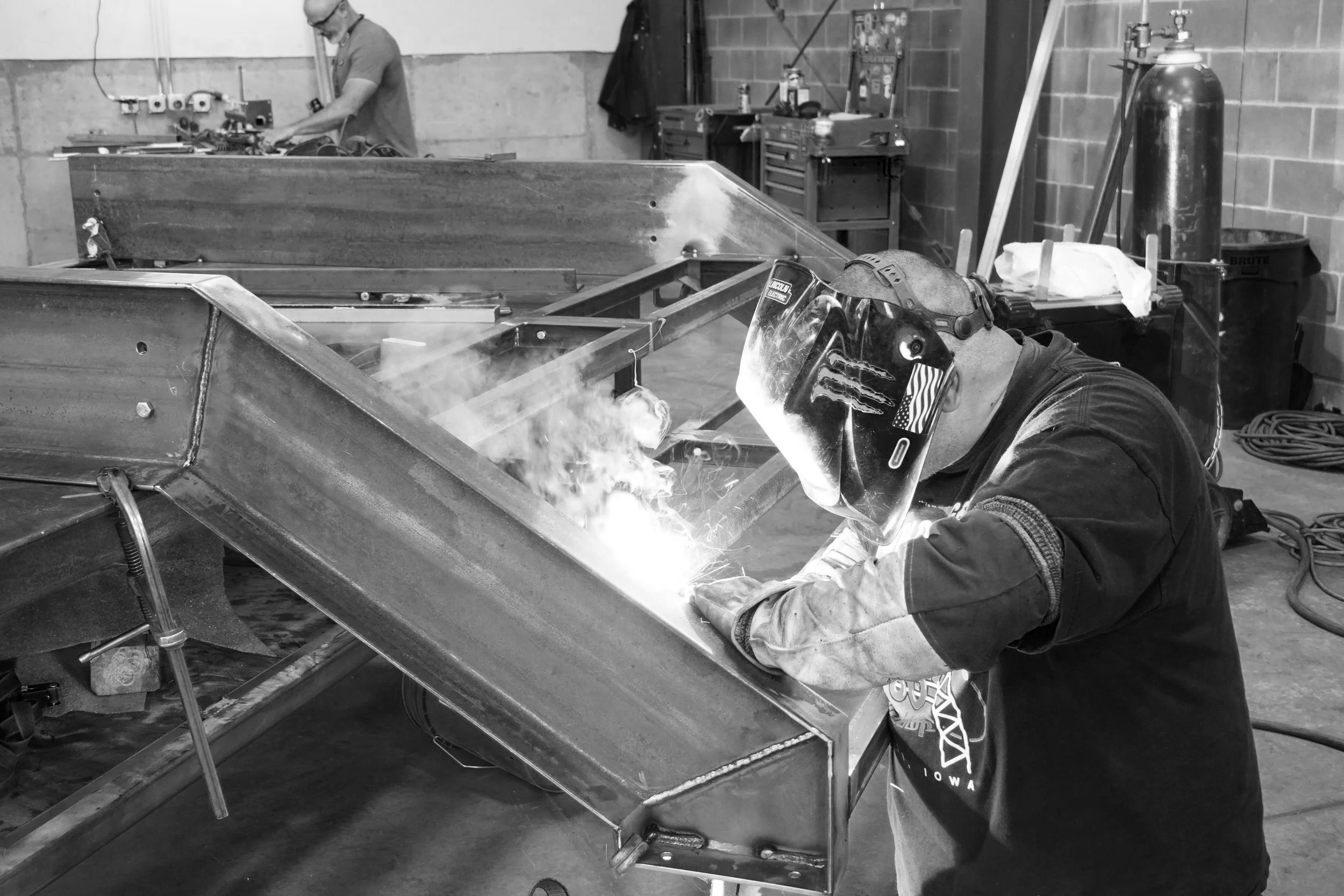 Person welding metal frame in a workshop, wearing a welding helmet and protective gloves.