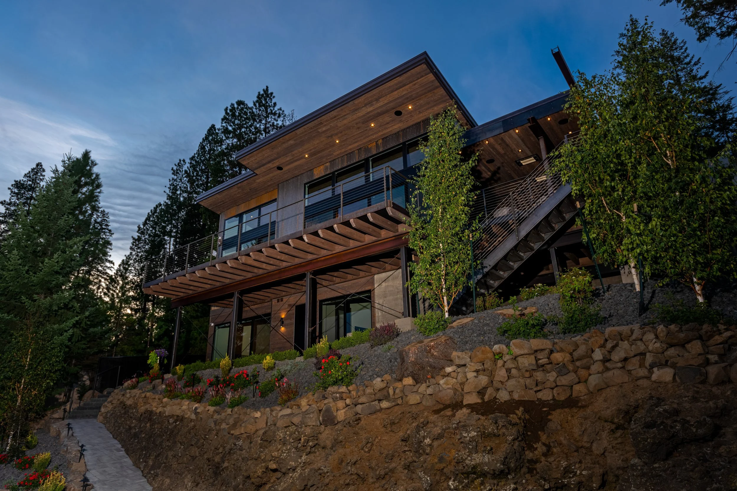 Modern multi-story house built into a hillside, with large glass windows and a wooden and metal exterior, surrounded by trees and a landscaped garden.