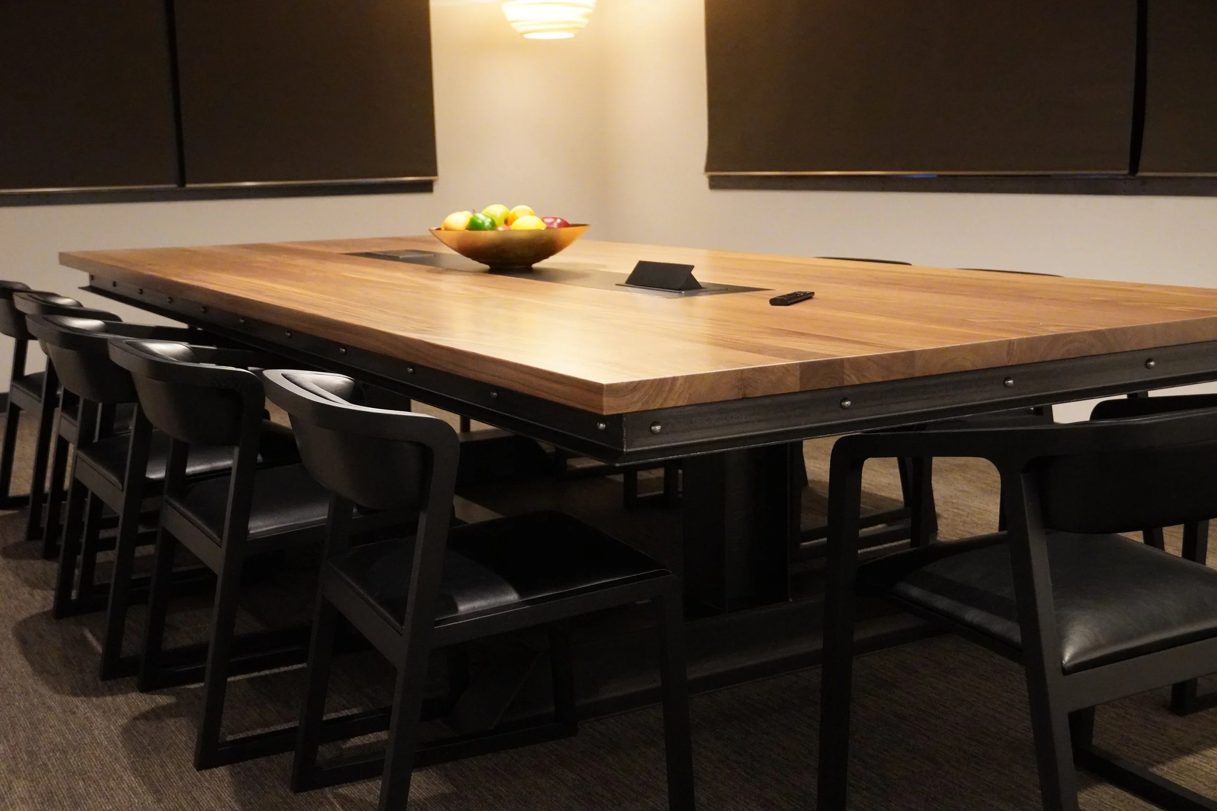 A large wooden conference table with black chairs around it, a bowl of colorful apples in the center, a remote, and a small black sign on the table. There are blackboards on the wall behind the table and a ceiling light.