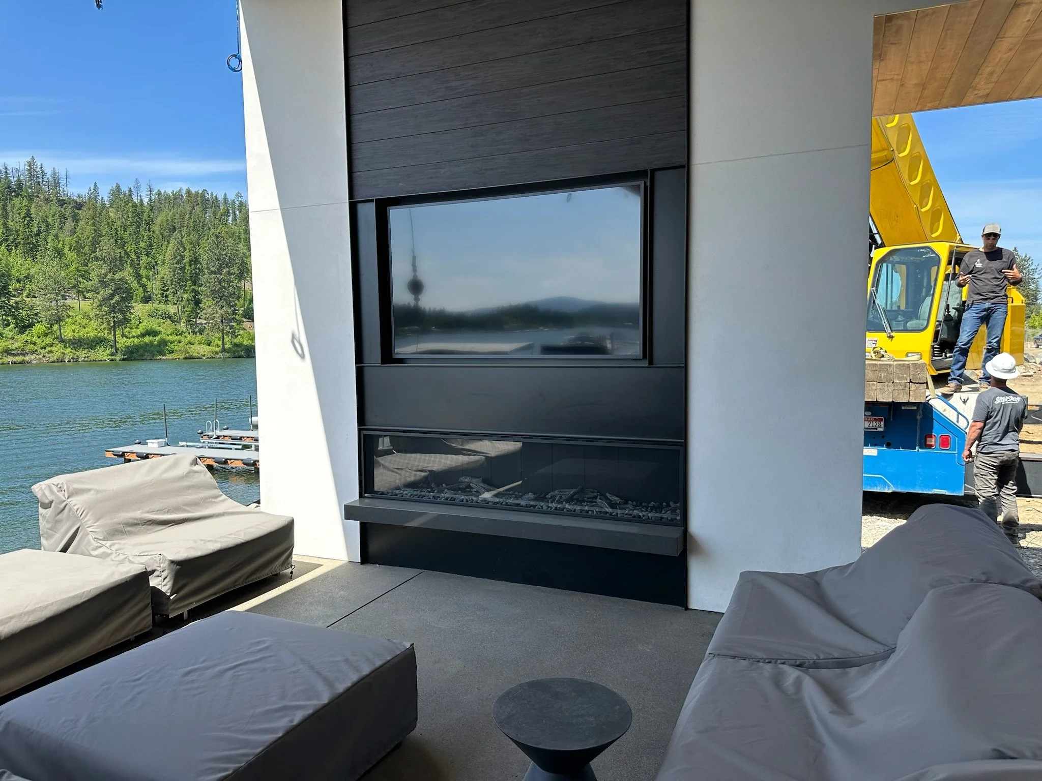 Modern living room with a fireplace, beige outdoor furniture, and a view of a lake and trees outside. Construction workers and a crane are visible on the right side of the image.